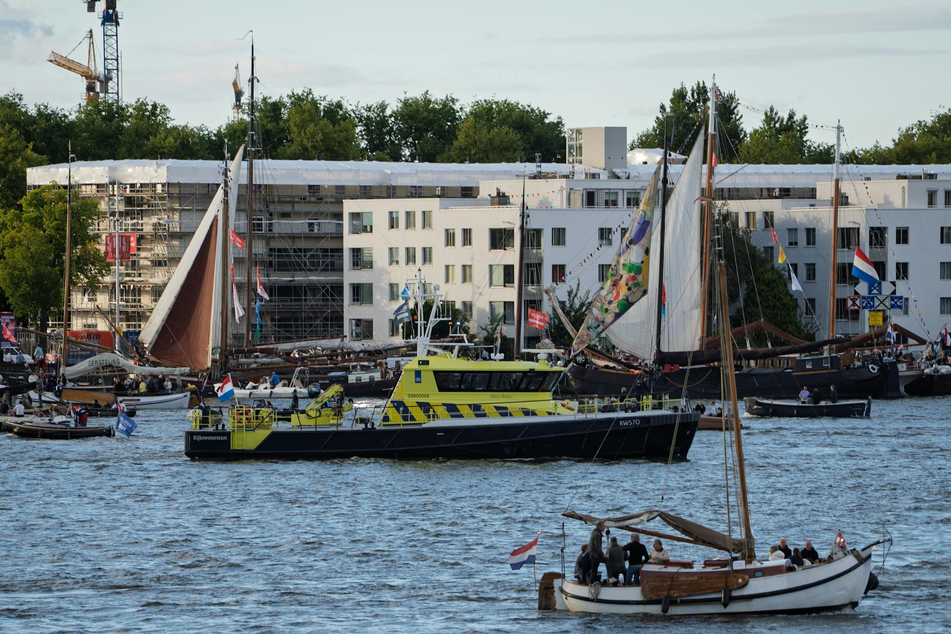 Colorful boats and vibrant sails navigate a bustling Amsterdam canal during a lively maritime event.
