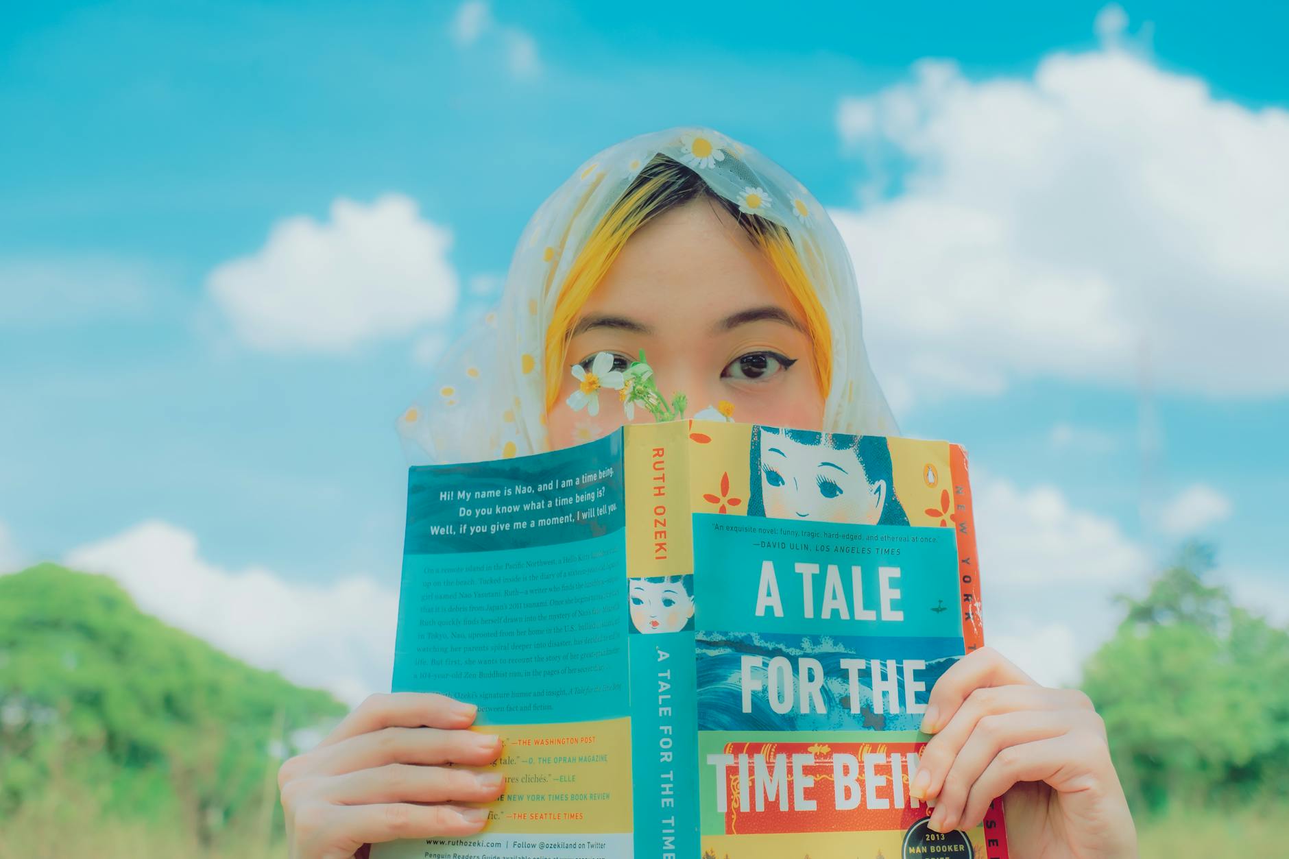 A woman with a headscarf reads 'A Tale for the Time Being' under a blue sky.