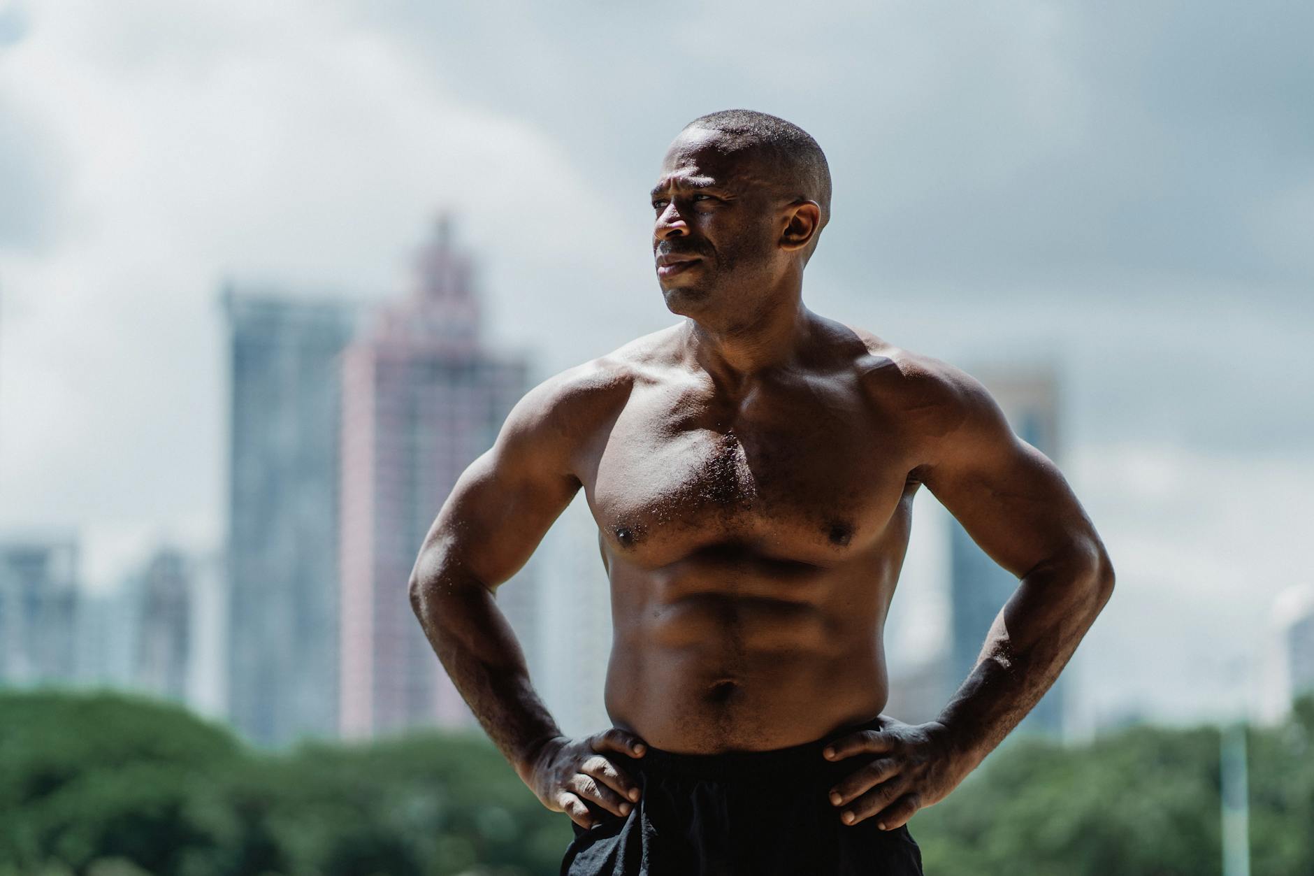 Muscular man posing shirtless outdoors against a city skyline, showcasing strength and vitality.