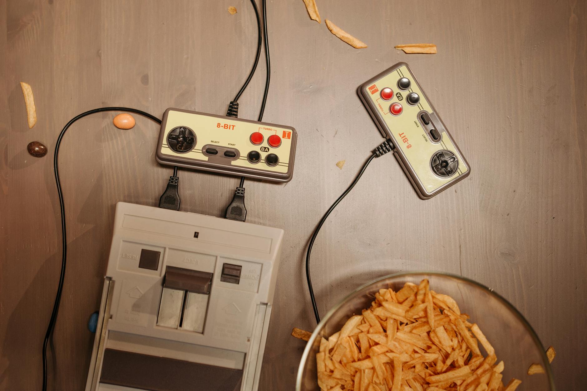 Top view of a retro gaming setup with controllers, console, and snacks on a wooden surface.