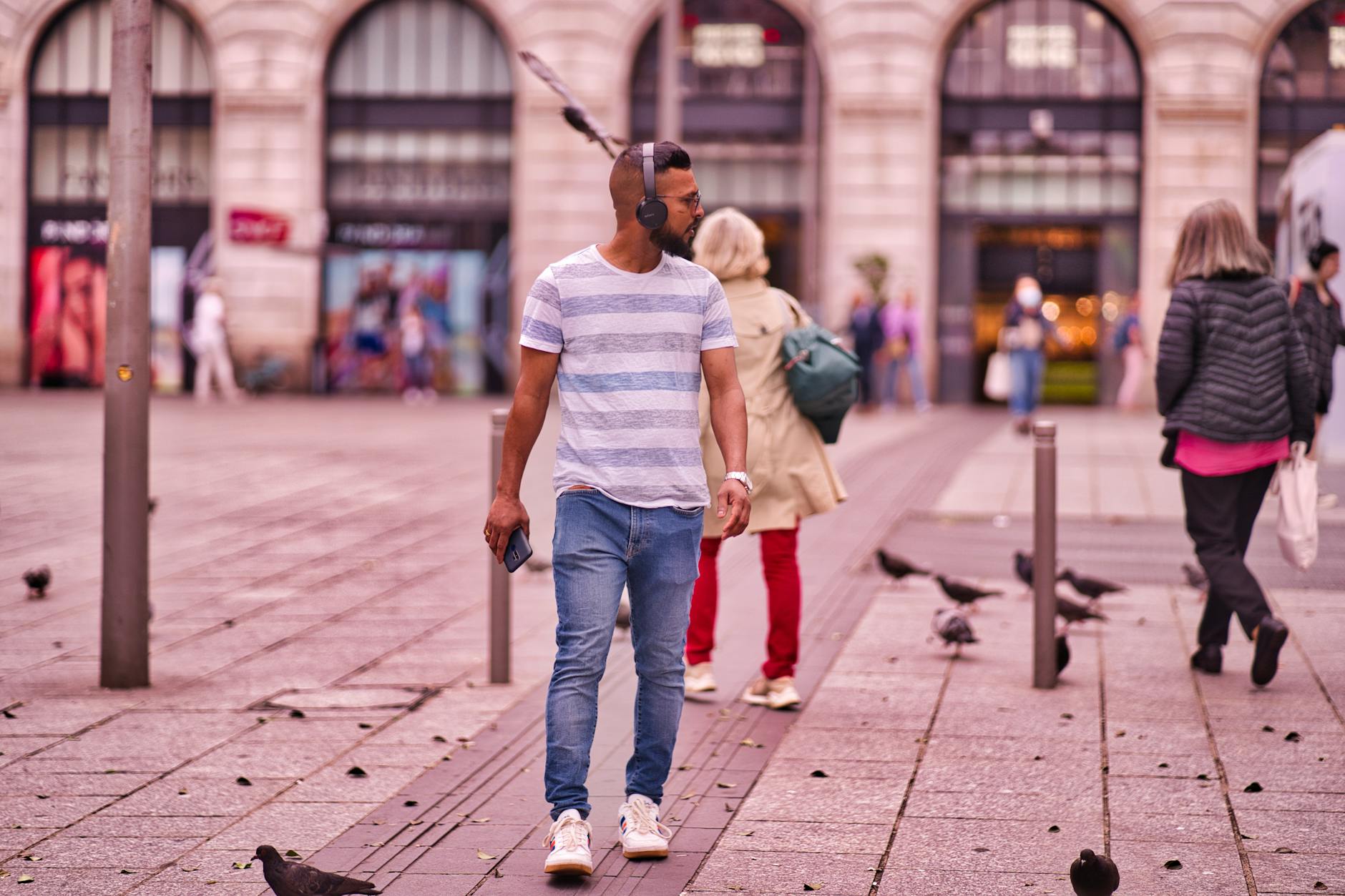 A man wearing headphones walks through a bustling city plaza surrounded by pigeons.