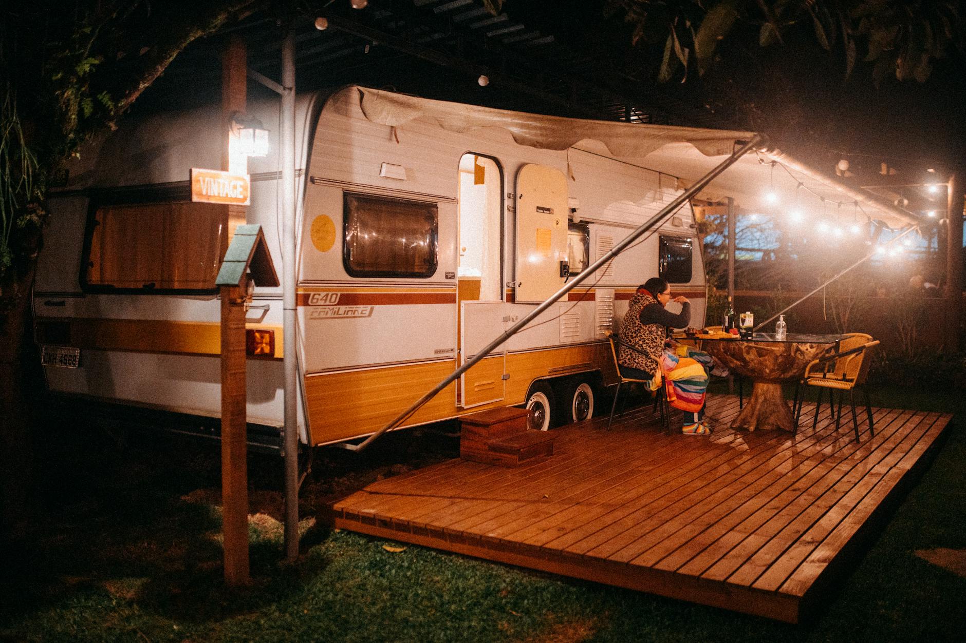 Side view of female tourist at table with alcoholic drink against camping trailer with shiny lamps at night
