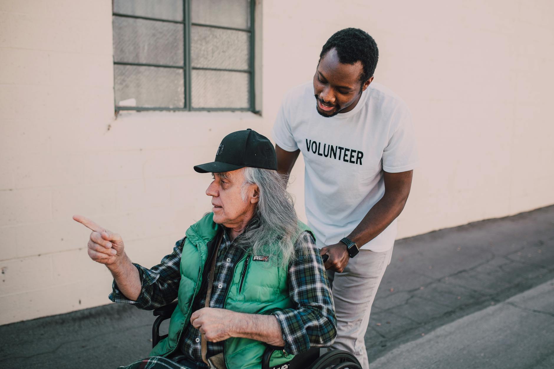 A volunteer helps an elderly man in a wheelchair outside, showing community support and care.