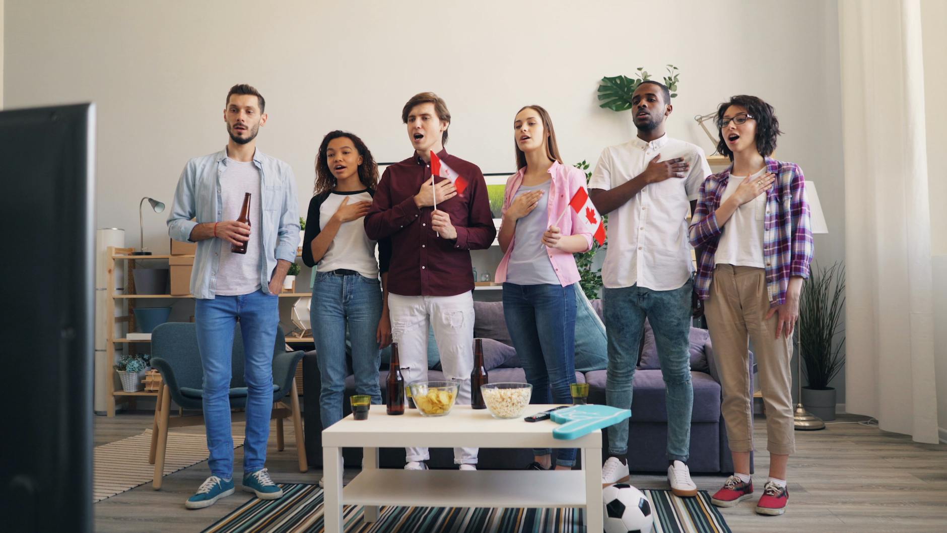 A diverse group of friends indoors, singing along to the national anthem while watching a TV broadcast.