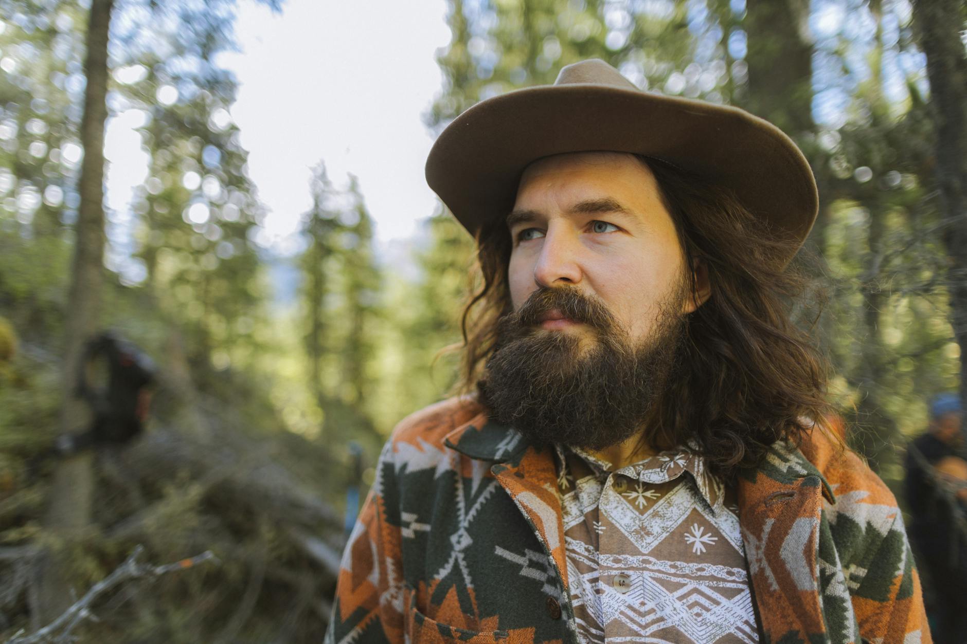 A bearded man in a patterned jacket and hat stands in a sunlit forest.