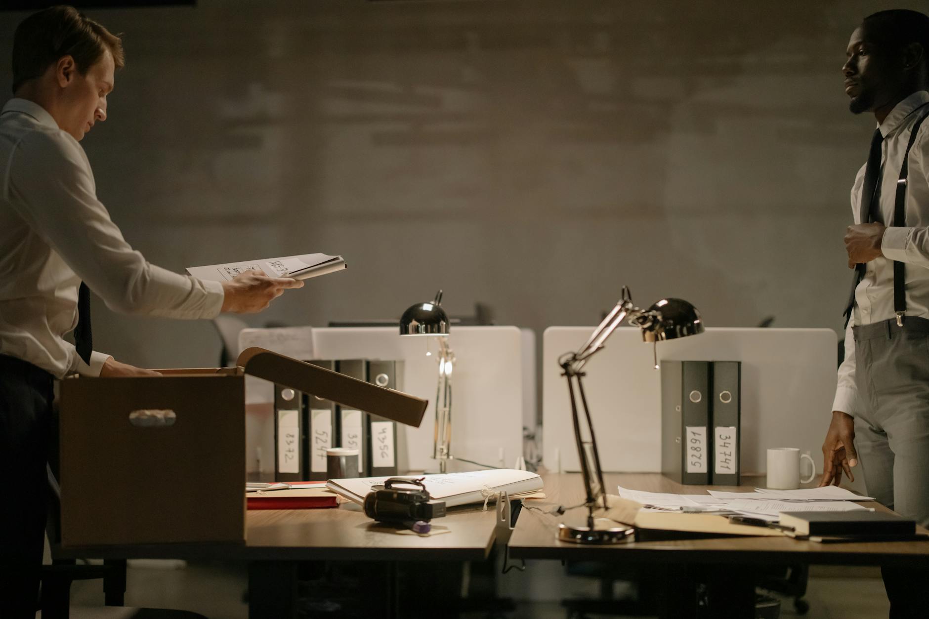 Two office workers sorting documents in a dimly lit workspace with several file binders.
