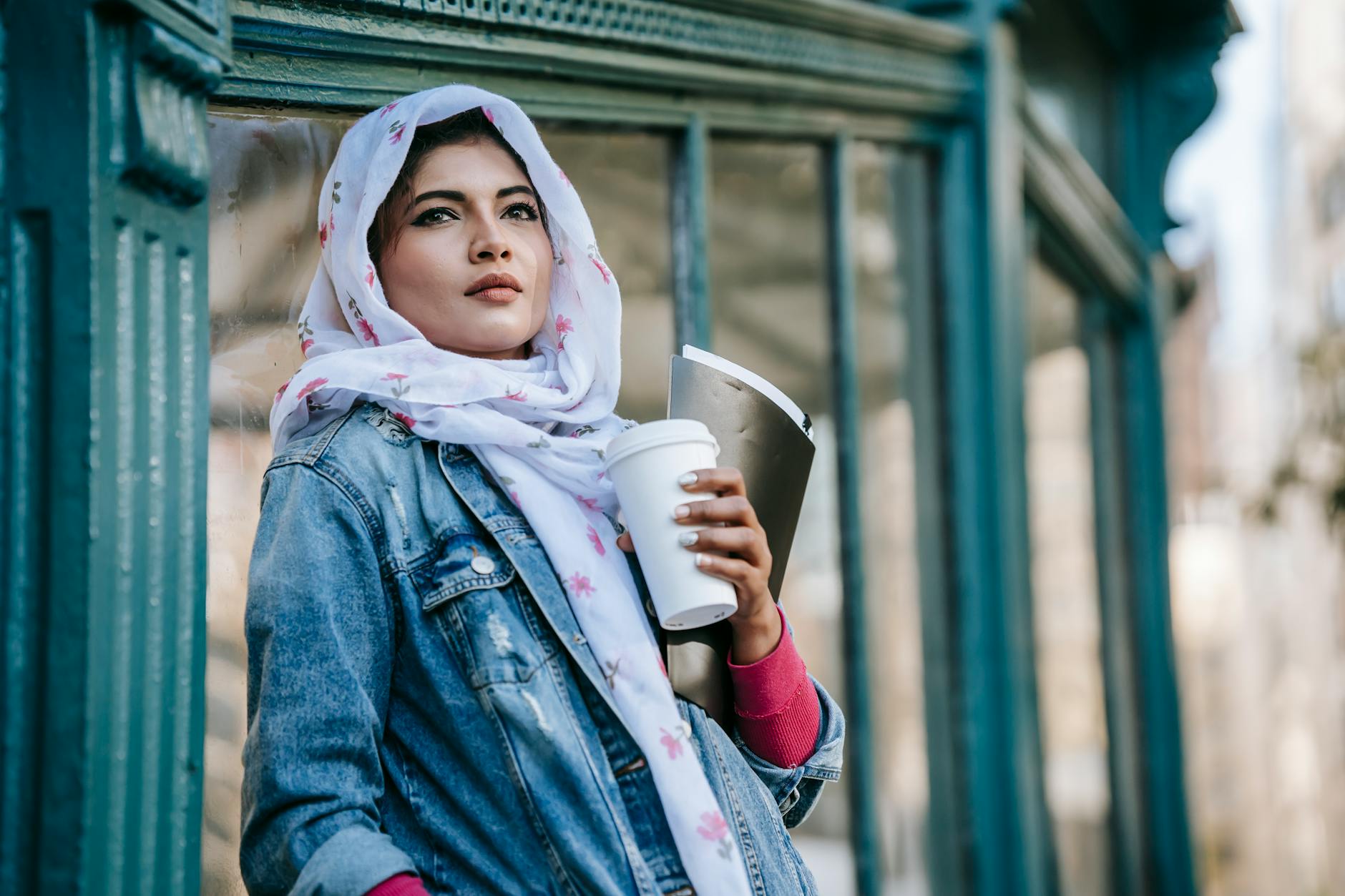 Stylish young woman in hijab holding a coffee outdoors, embracing a contemporary urban lifestyle.