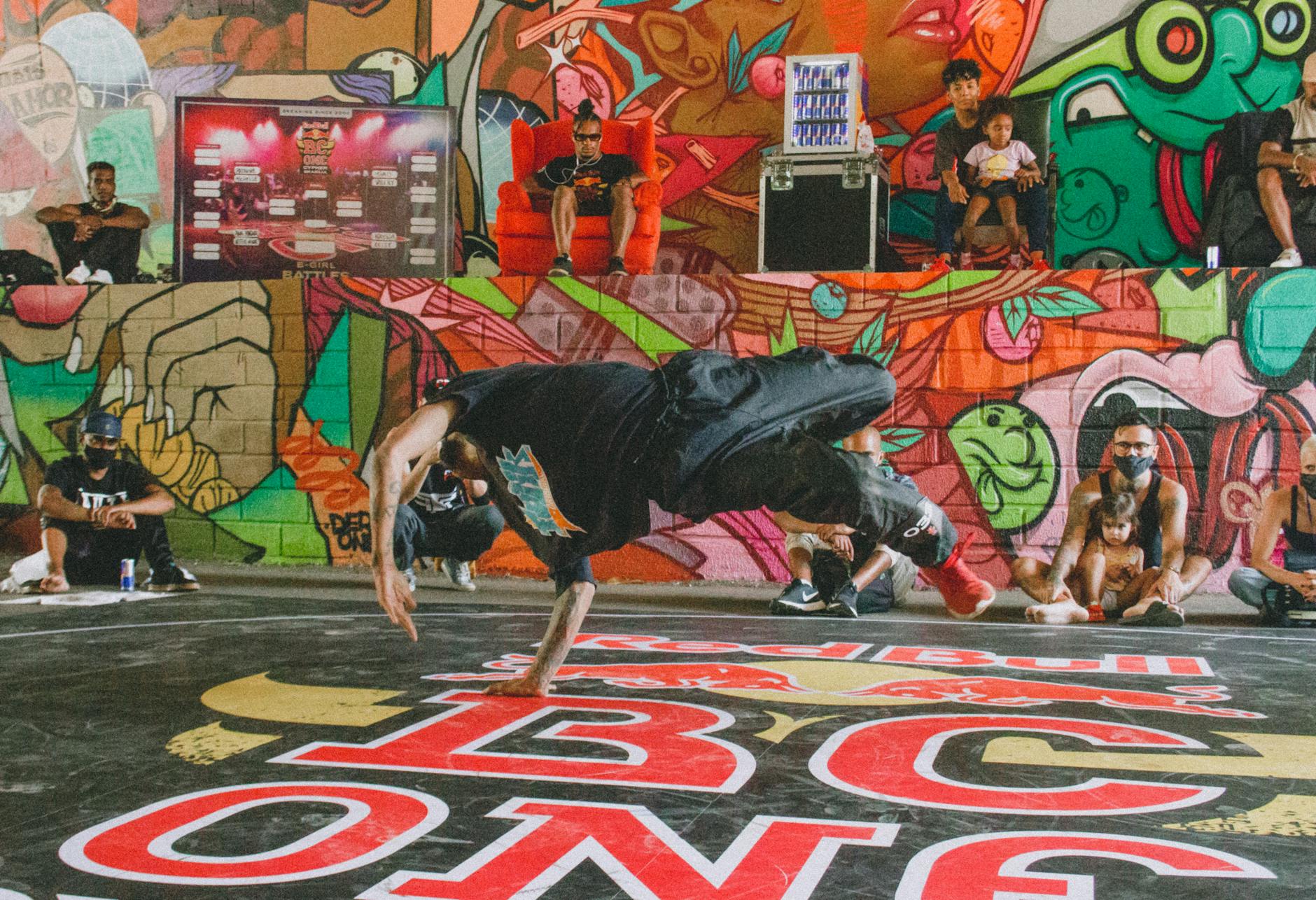 Energetic breakdancer in action with vibrant graffiti and audience backdrop.