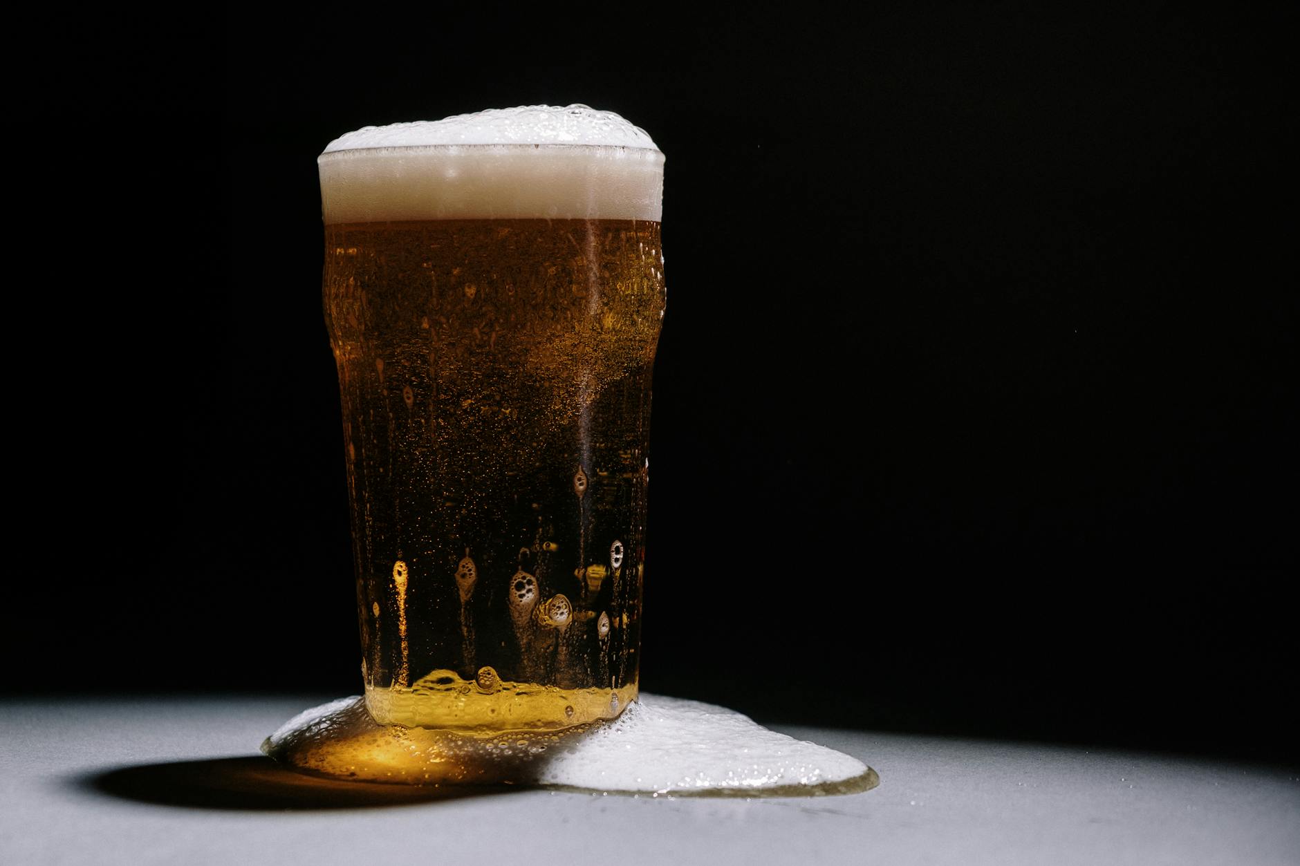 A studio shot of a frothy and overflowing pint of golden beer against a dark backdrop.