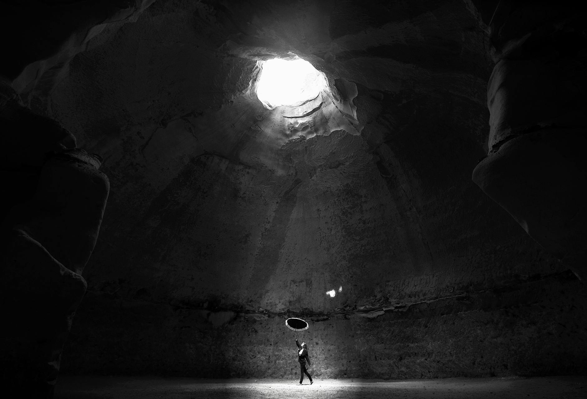 Black and white image of a person with an umbrella in a dramatic cave setting with natural skylight.