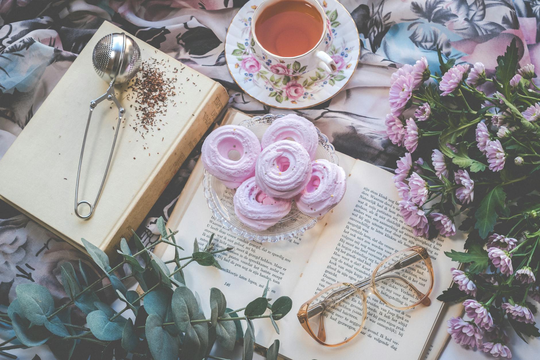 Delicate flatlay with pink pastries, tea, flowers, book, and glasses for a romantic setting.
