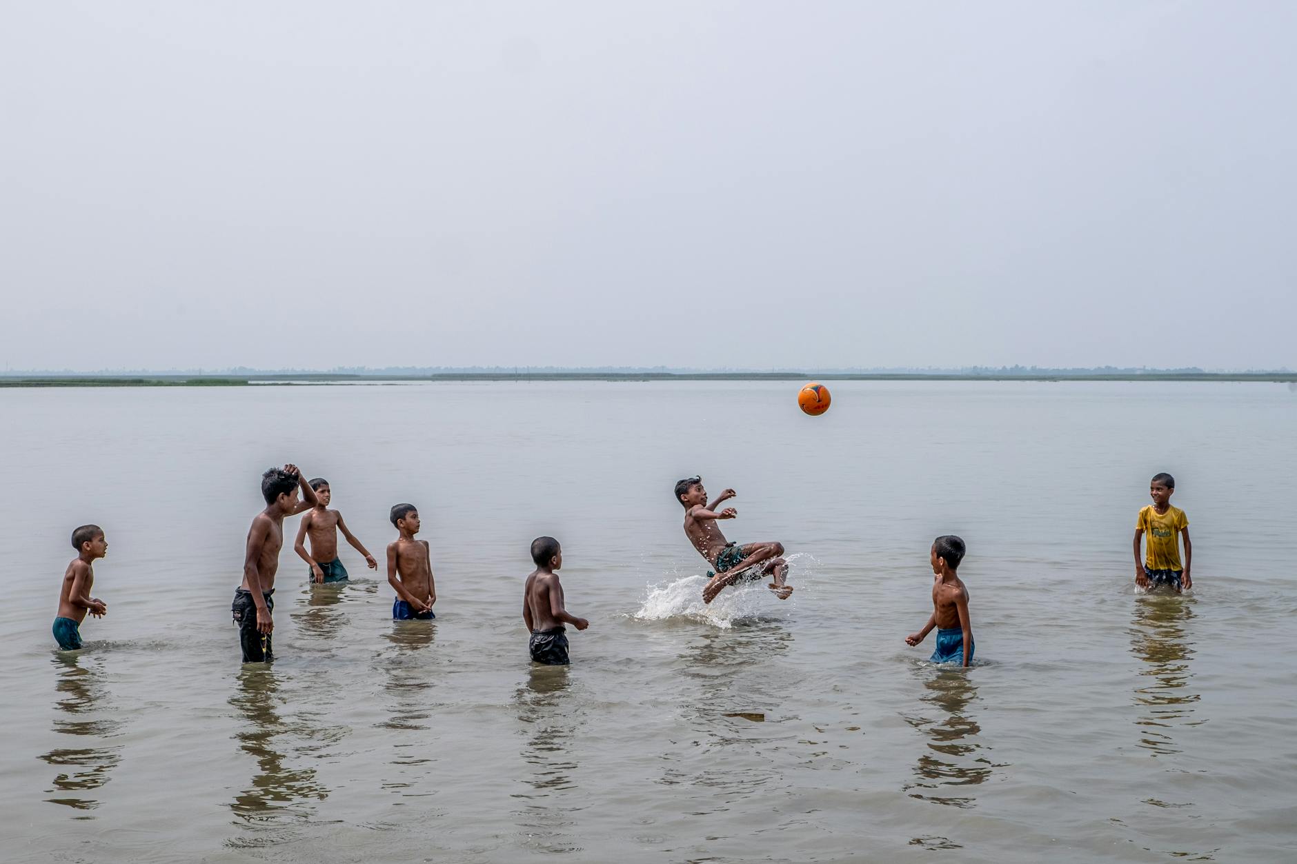 Joyful group of boys playing soccer in shallow water with an orange ball on a sunny day.