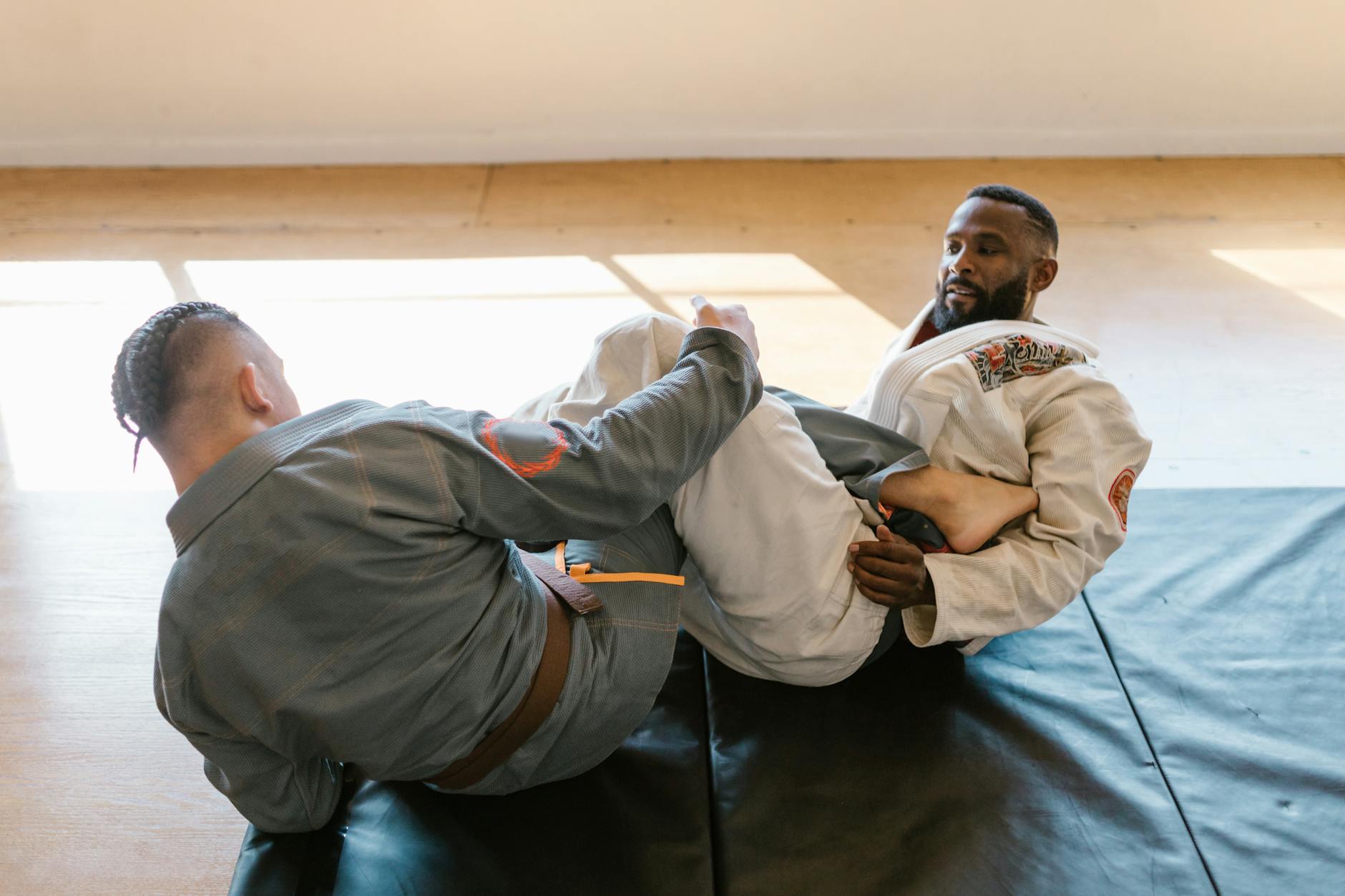 Two men in martial arts gis practicing Brazilian Jiu Jitsu indoors with natural light.
