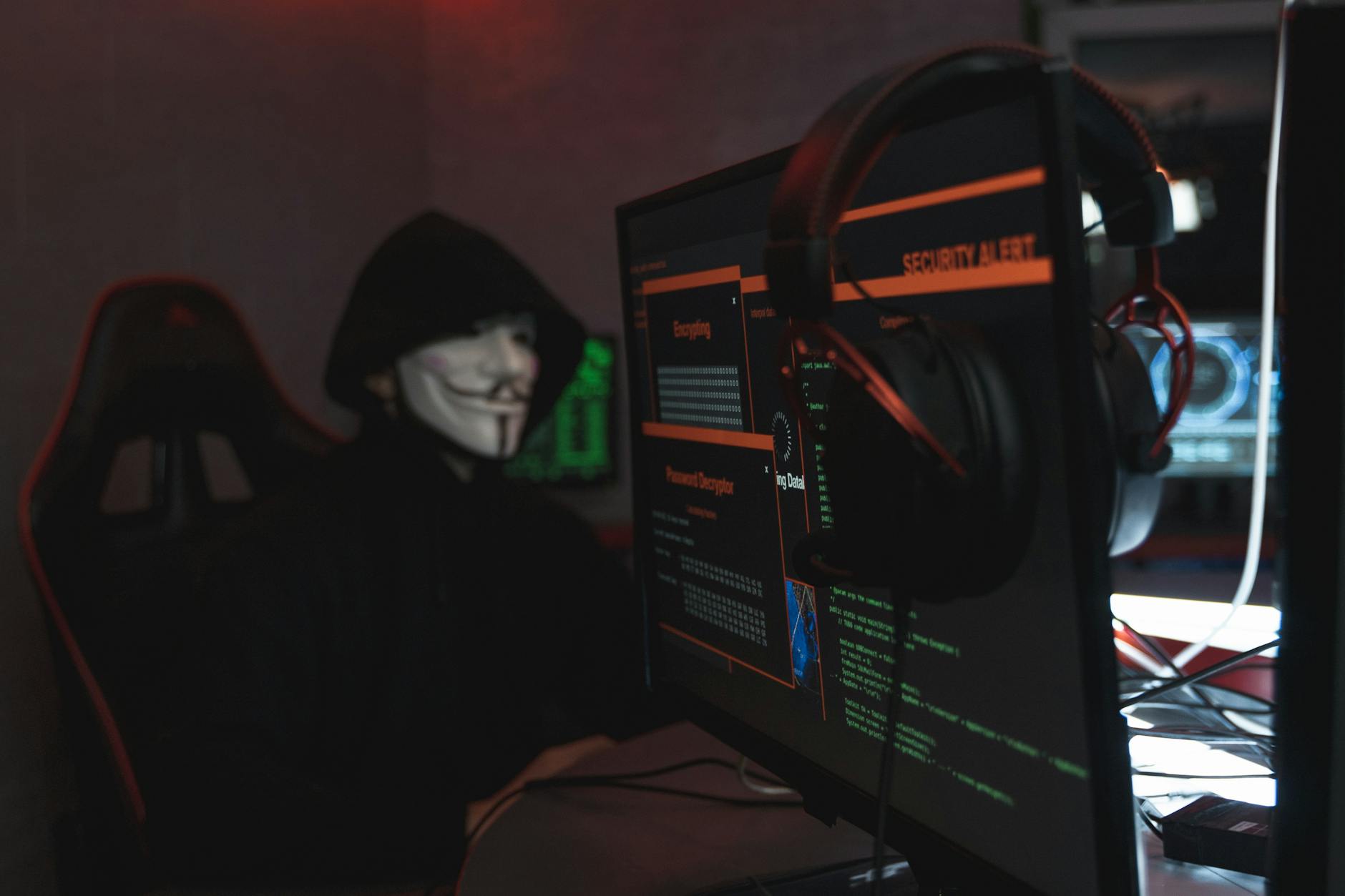A person wearing a Guy Fawkes mask engaged in hacking activities on a computer in a dimly lit room.