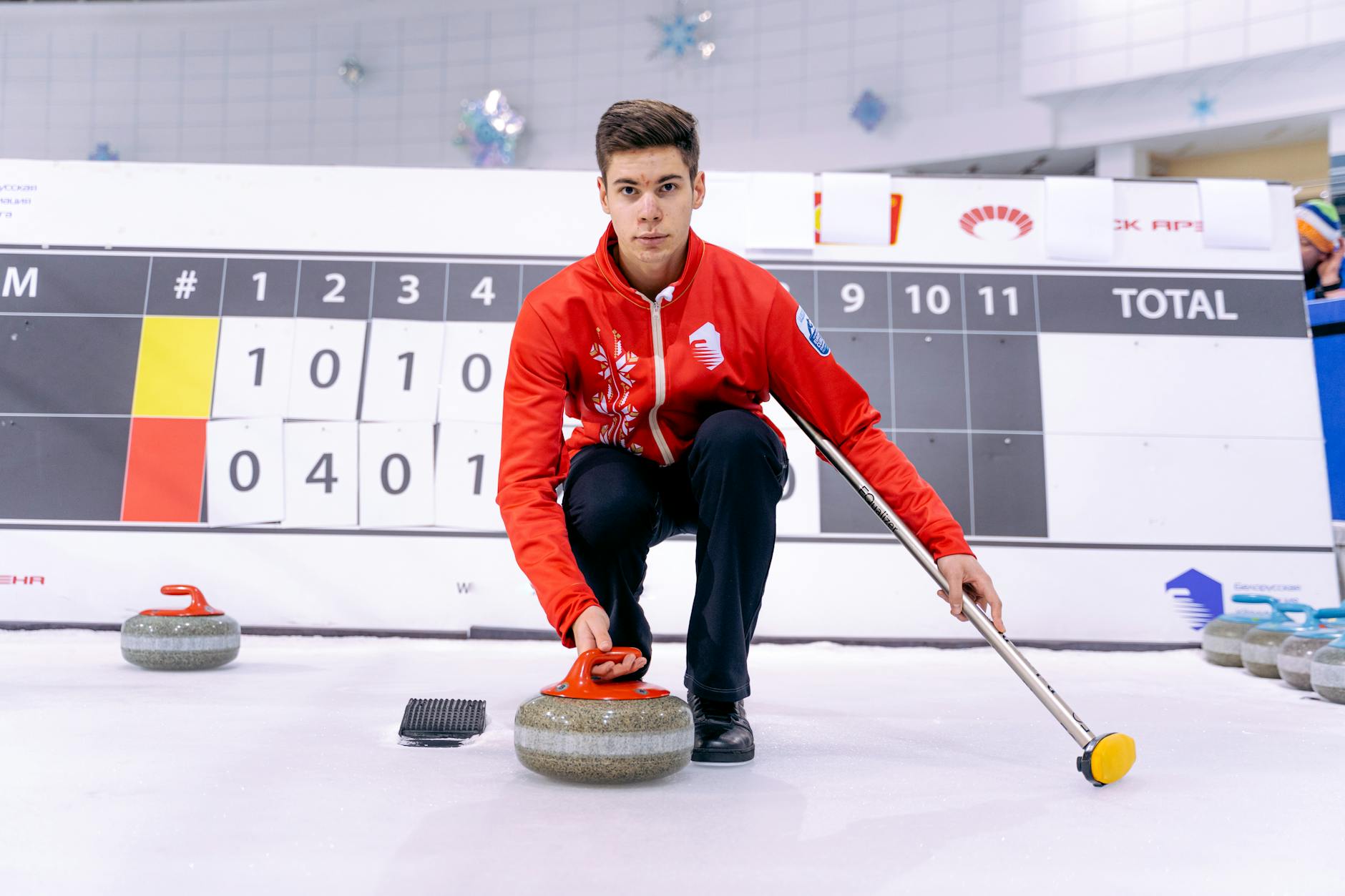 Young male athlete in a red jacket skillfully playing curling indoors on an ice rink.