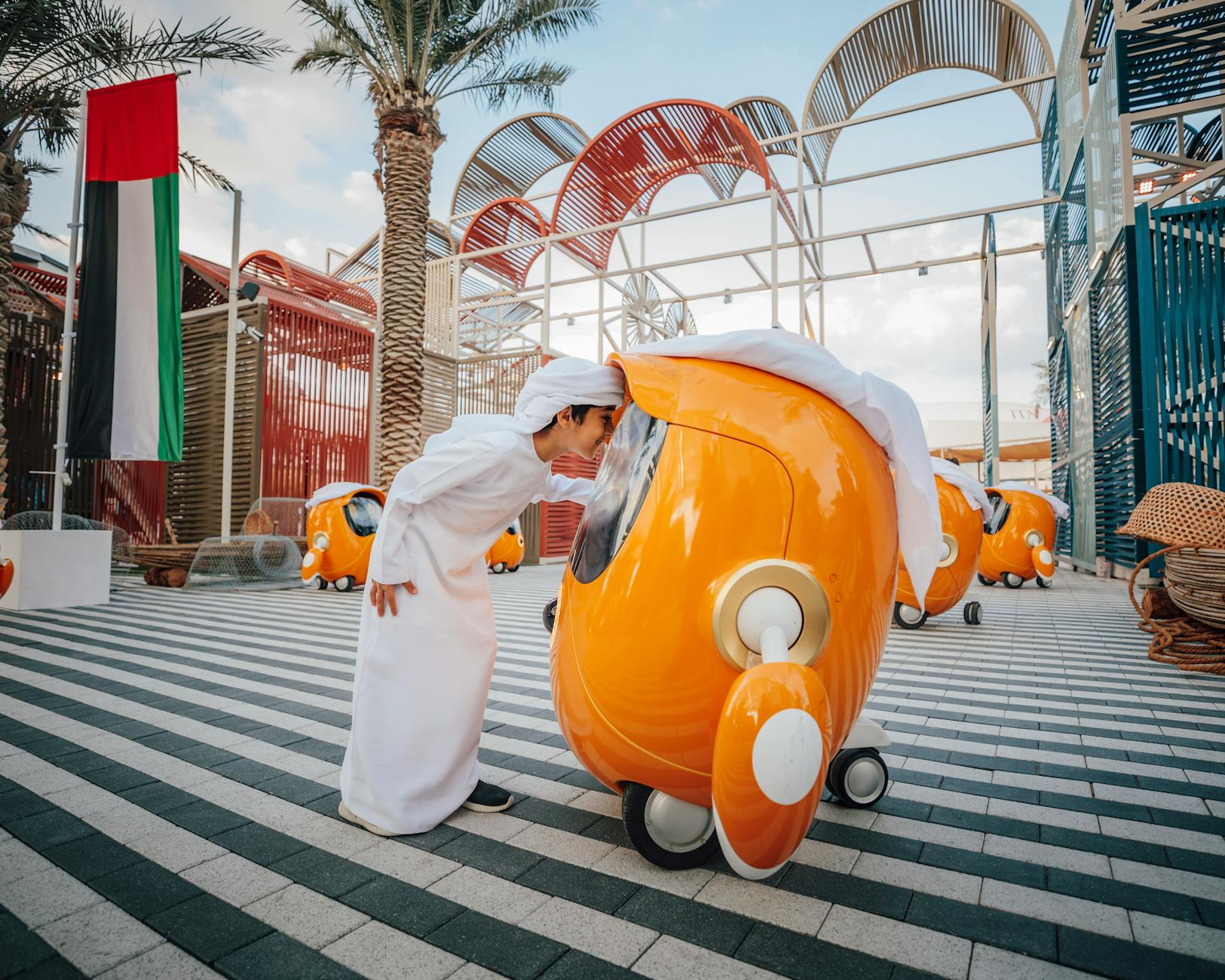 A child in traditional Arabic attire interacts with a futuristic orange robot in Dubai's urban setting.