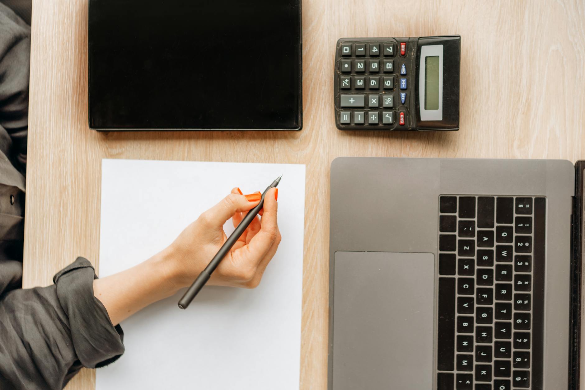A person writing on paper surrounded by a laptop, tablet, and calculator on an office desk.