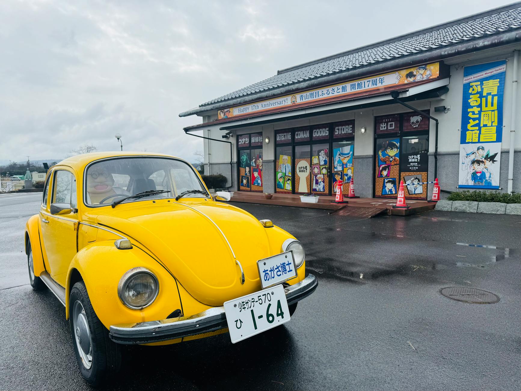 Yellow vintage car parked outside a Conan-themed store on a rainy day.
