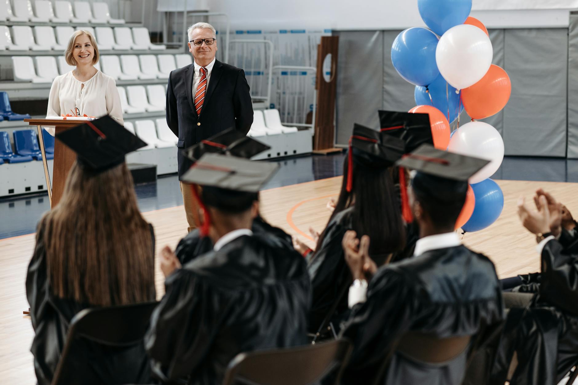 Graduation ceremony in a gymnasium with speakers addressing graduates seated in gowns.