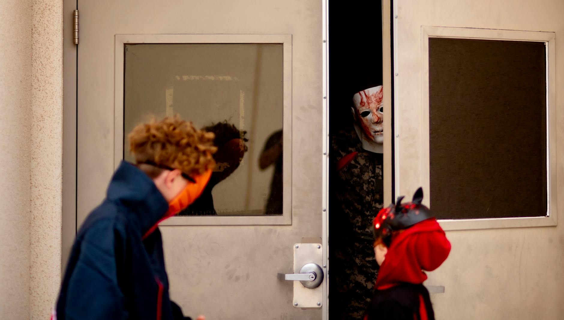 Children in Halloween costumes approach a spooky doorway with a figure in a mask peeking out.