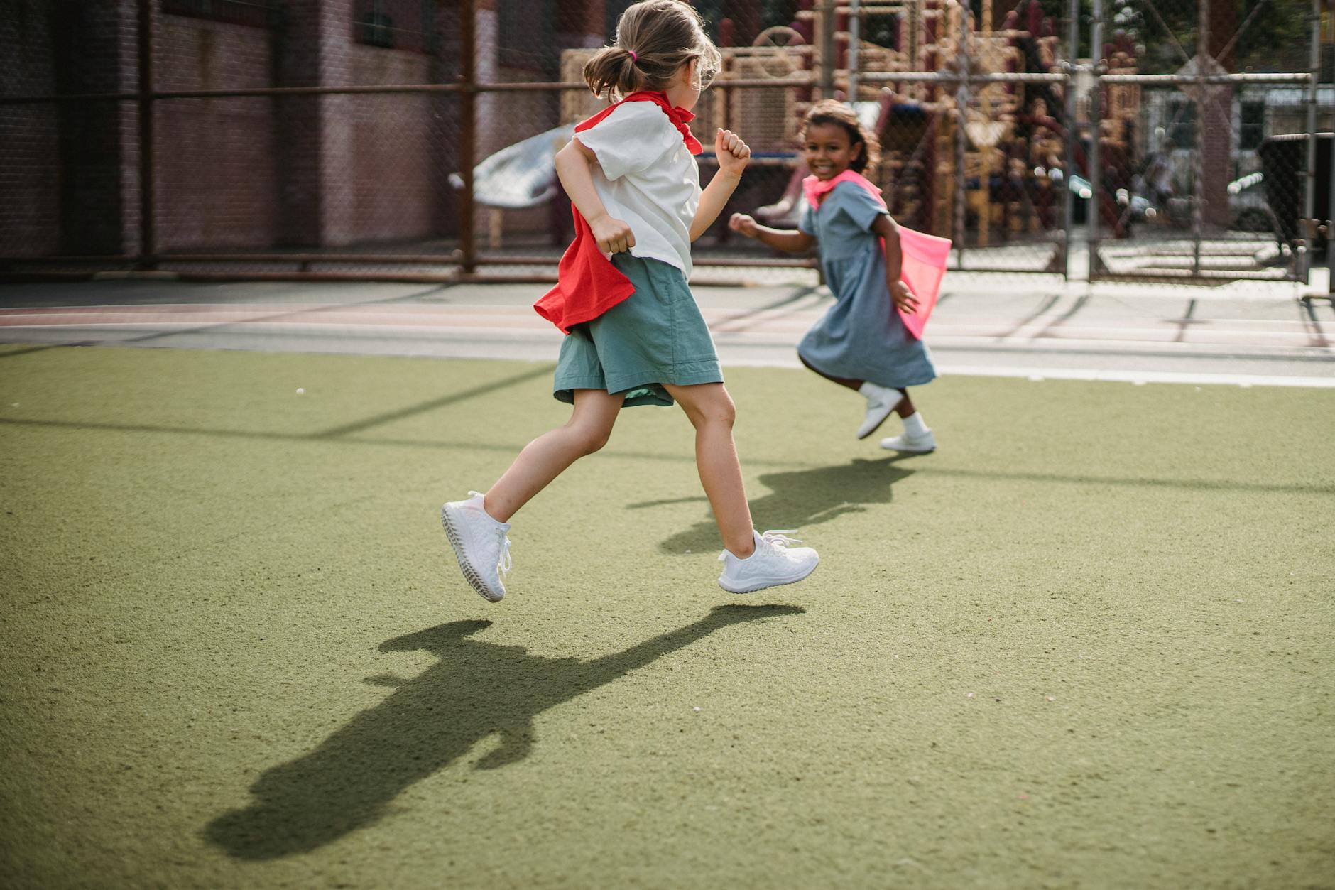 Two children happily running with capes, enjoying playtime on a sunny day outdoors.