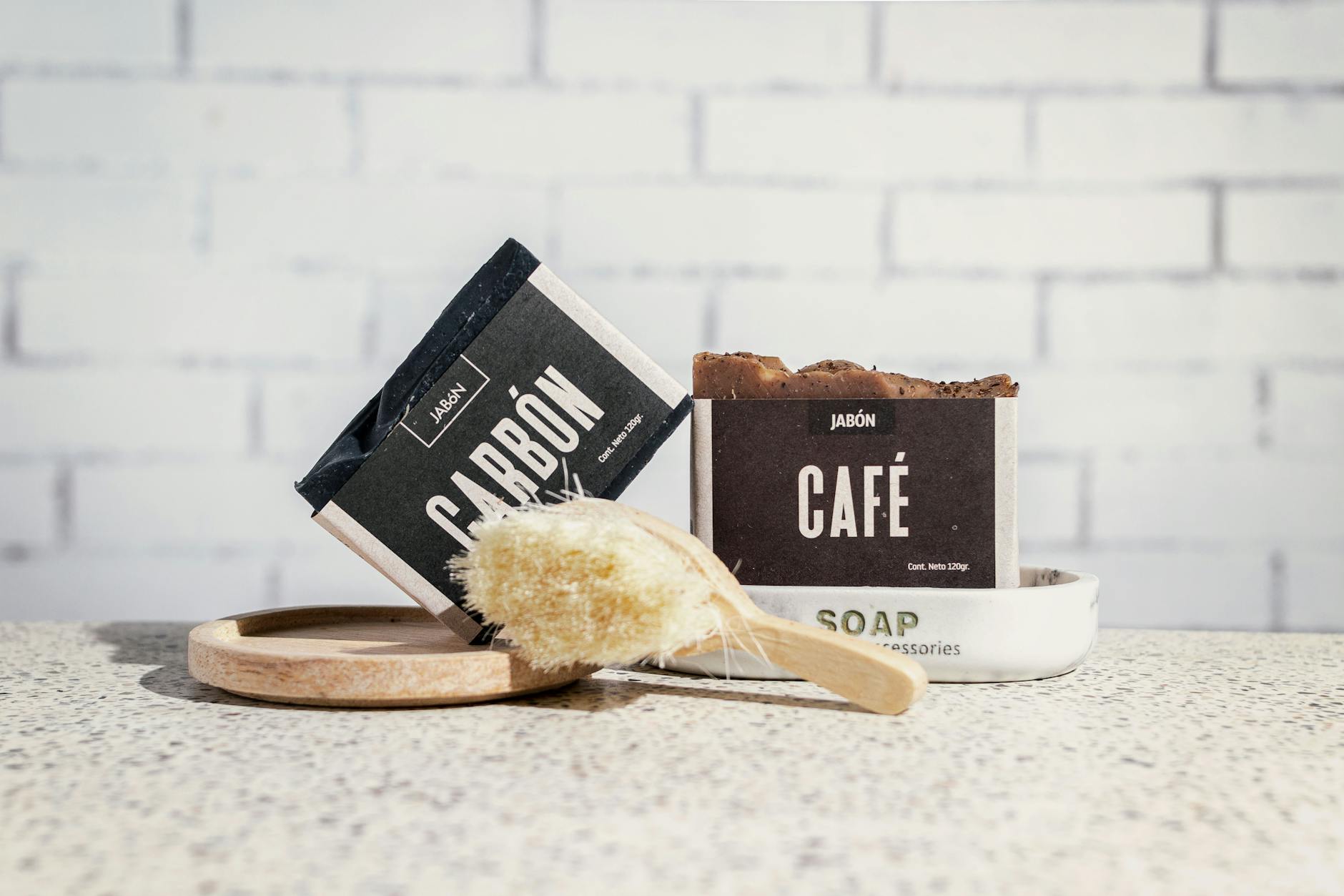 A close-up of artisanal coffee and charcoal soaps with a bath brush against a white brick wall.