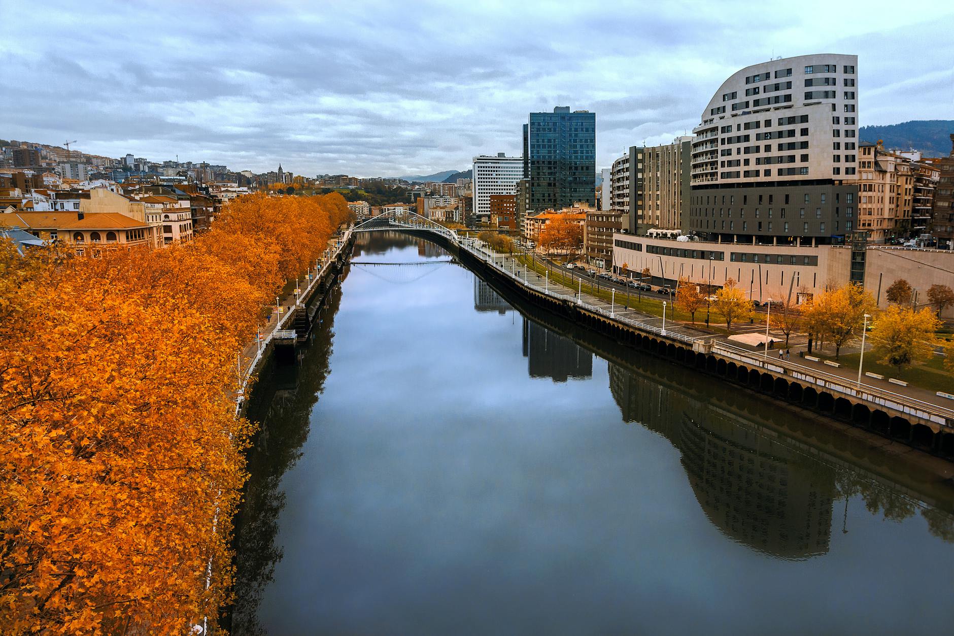 Autumn view of a river in Bilbao, Spain with orange foliage and modern skyline.