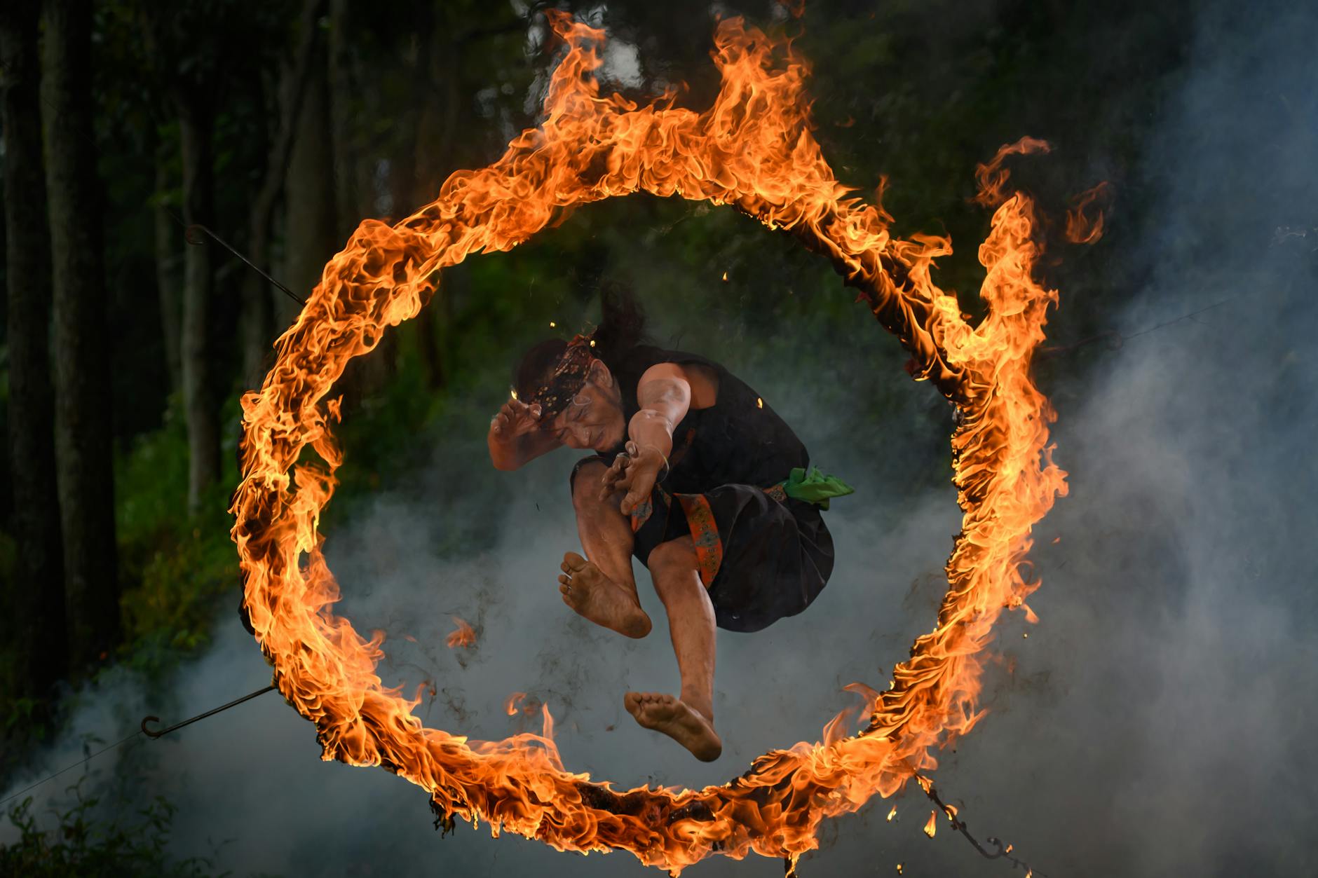 Daring man jumps through a flaming hoop in an outdoor forest setting.