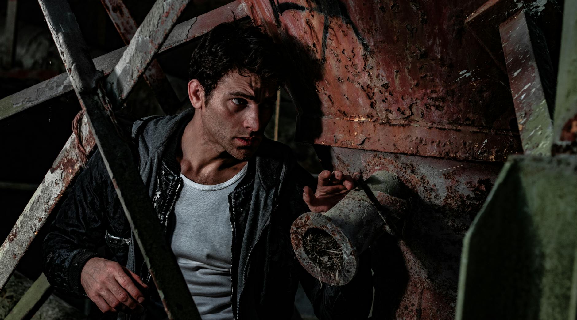Man hidden in a dark industrial setting, appearing cautious and tense in a rusty environment.