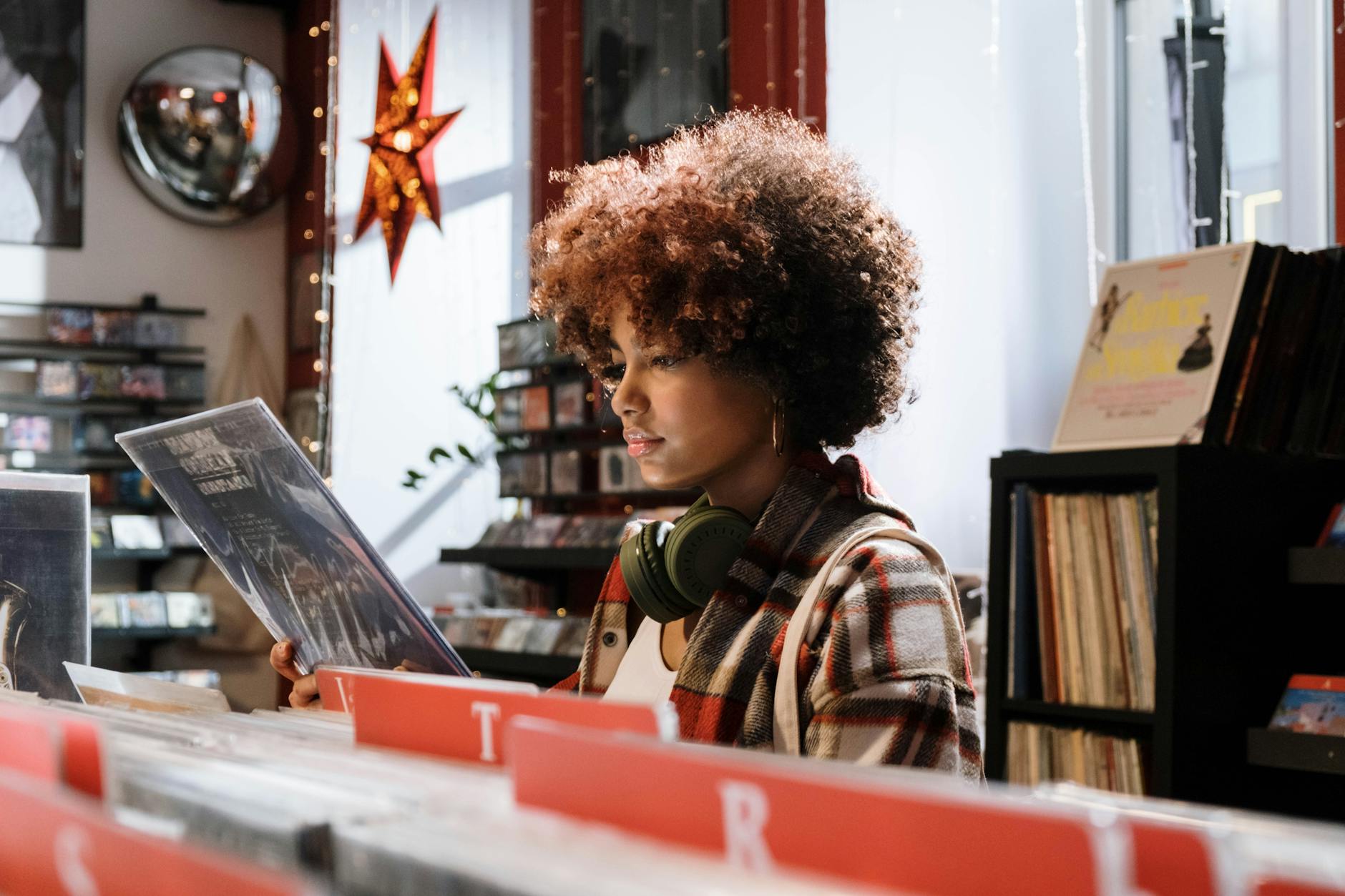A woman with afro hair examines vinyl records in a cozy music shop, wearing headphones.