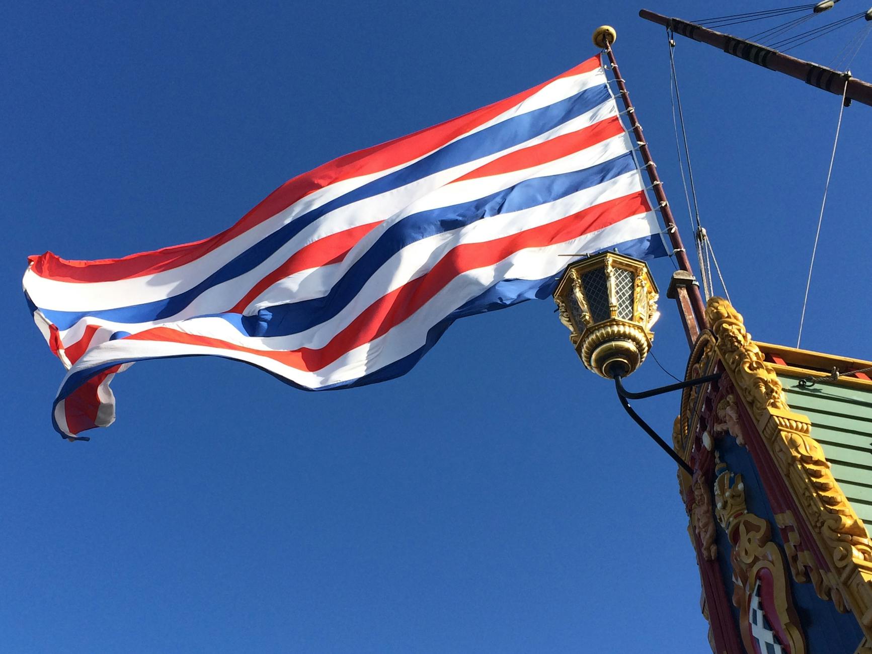 Vibrant red, white, and blue flag on an ornate ship under clear blue sky.