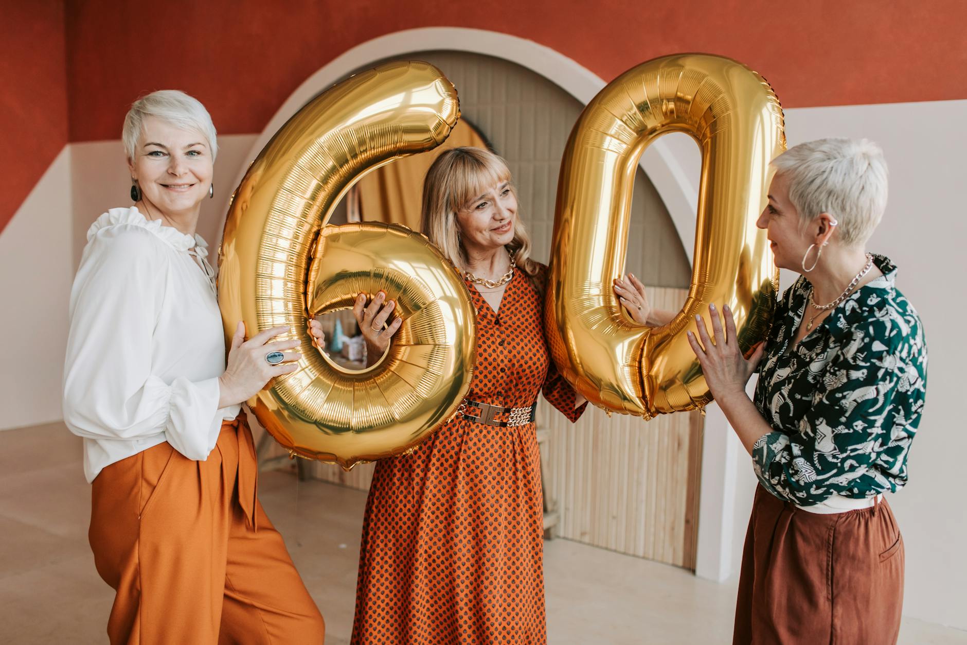 Three senior women joyfully celebrating a 60th birthday with golden balloons indoors.