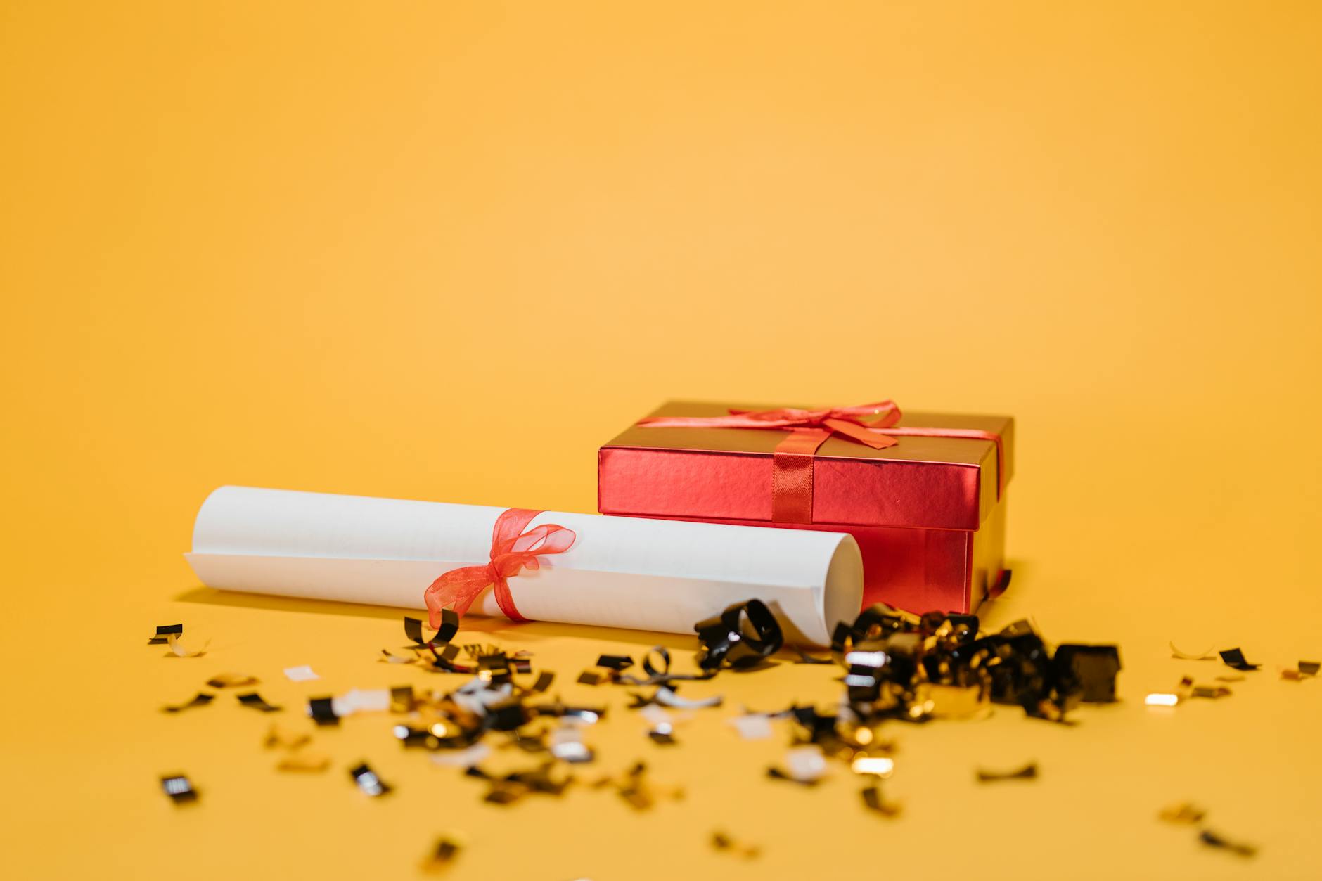 Celebratory image of a diploma and gift box amidst confetti on a vibrant yellow background.