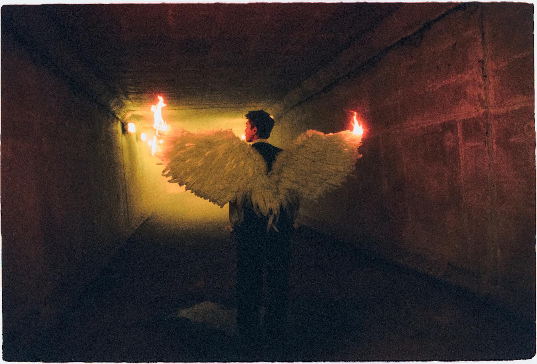 A person in angel wings holding fire walks through a moody, dimly lit tunnel.