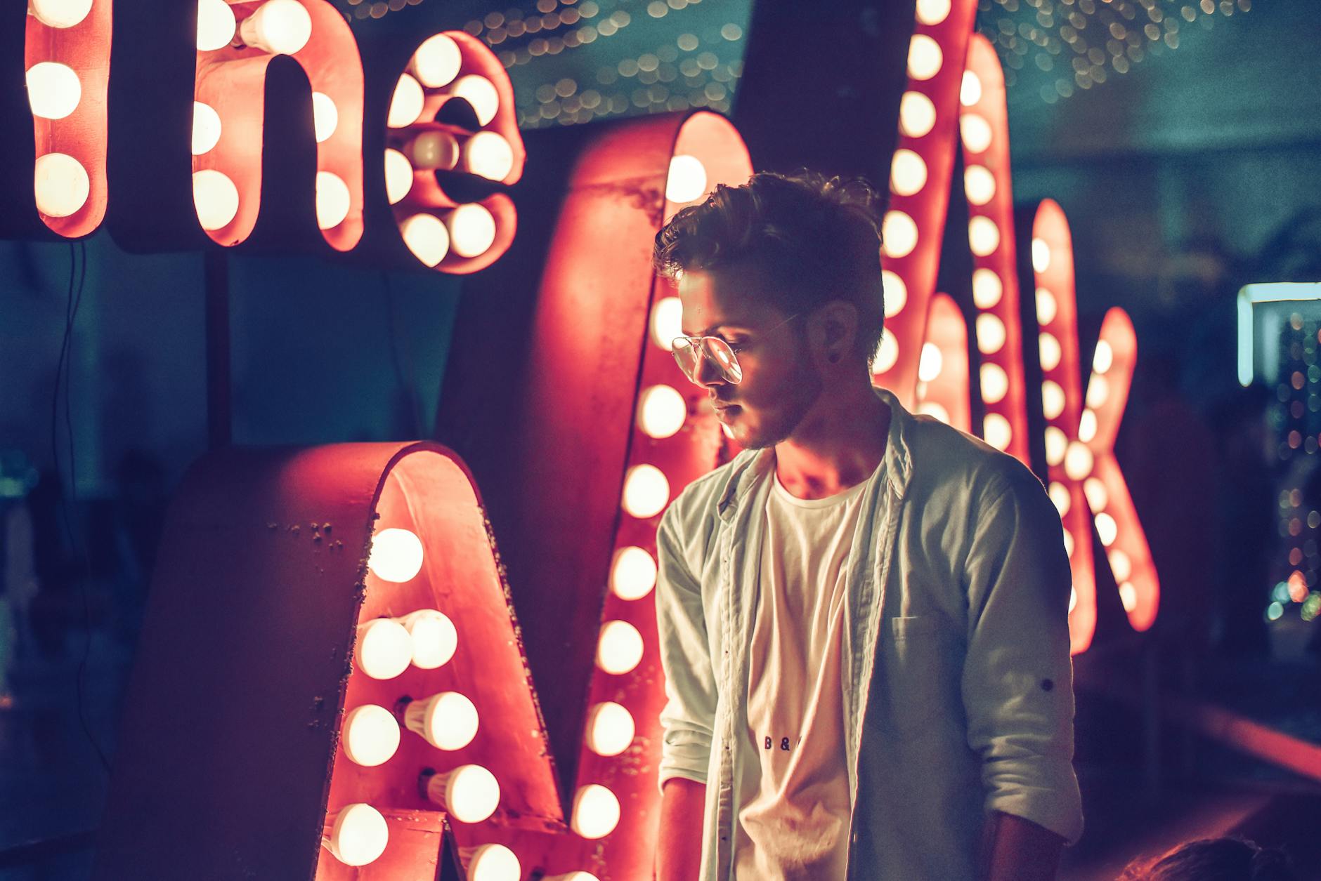 A stylish man stands near illuminated lettering in a vibrant nightclub setting, exuding a sense of nightlife glamour.