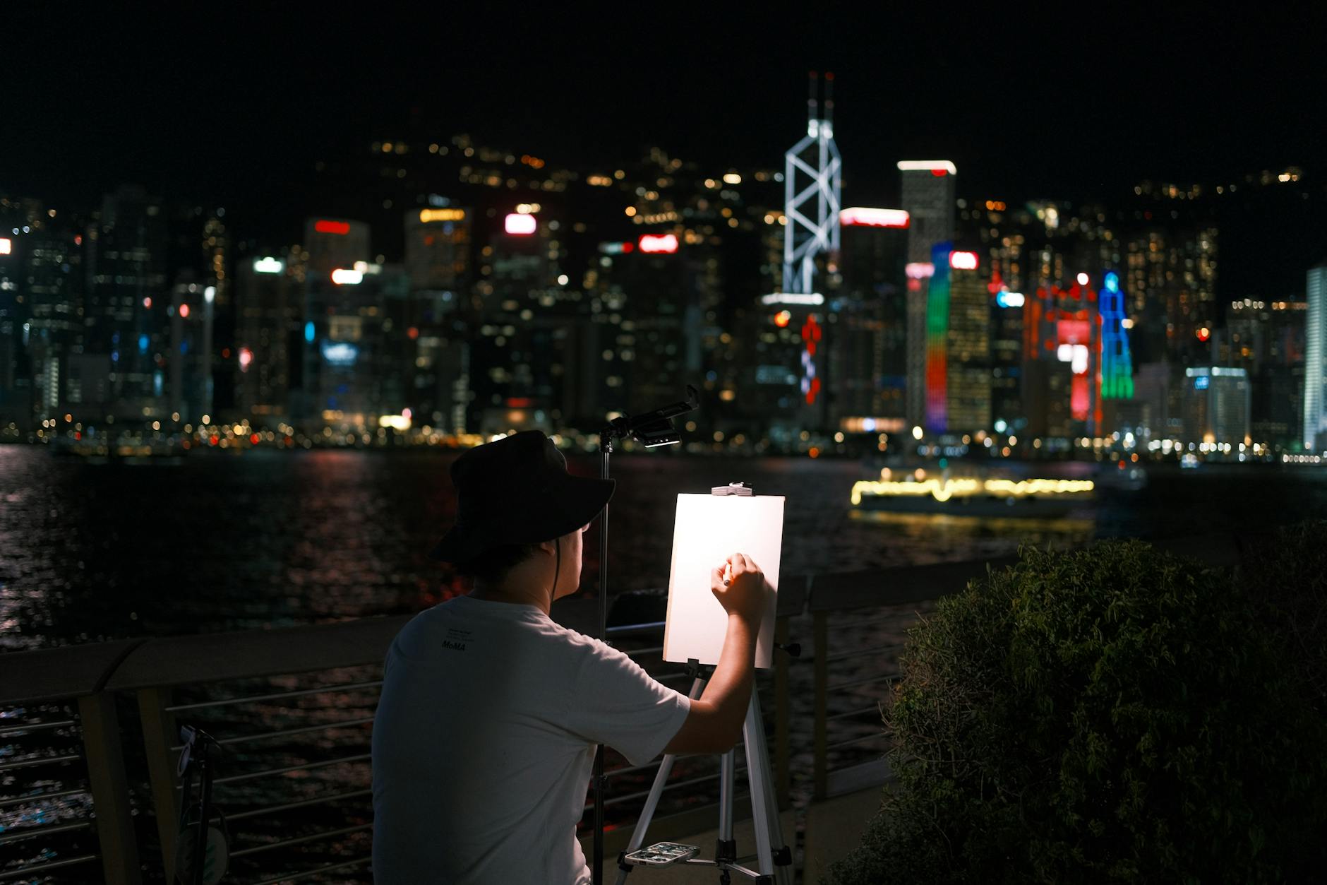 Artist sketching at night with vibrant Hong Kong cityscape as backdrop.