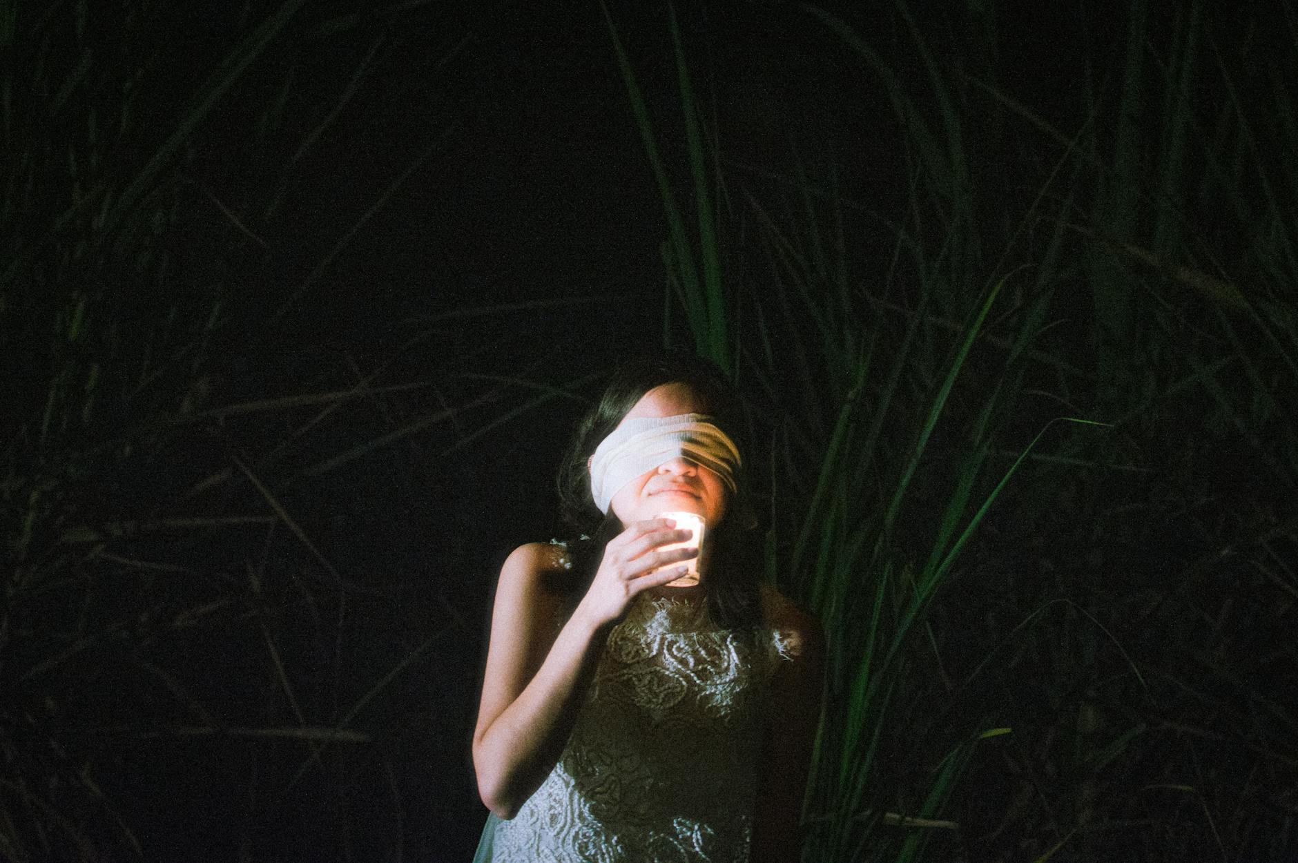 A blindfolded woman holds a candle in the dark, surrounded by vegetation in Veracruz, Mexico.
