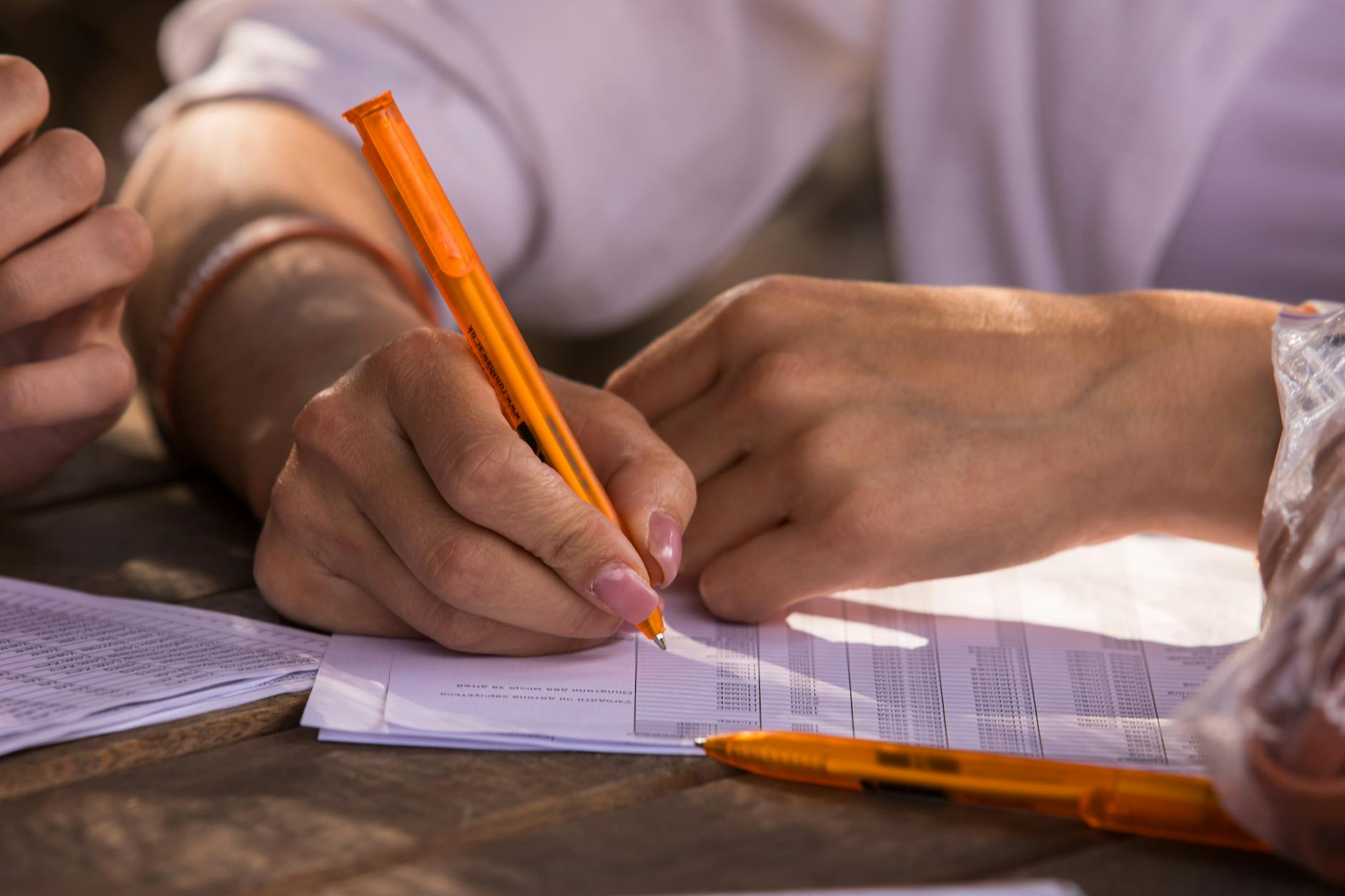 Close-up shot of hands writing on documents with orange pen, implying business or education setting.