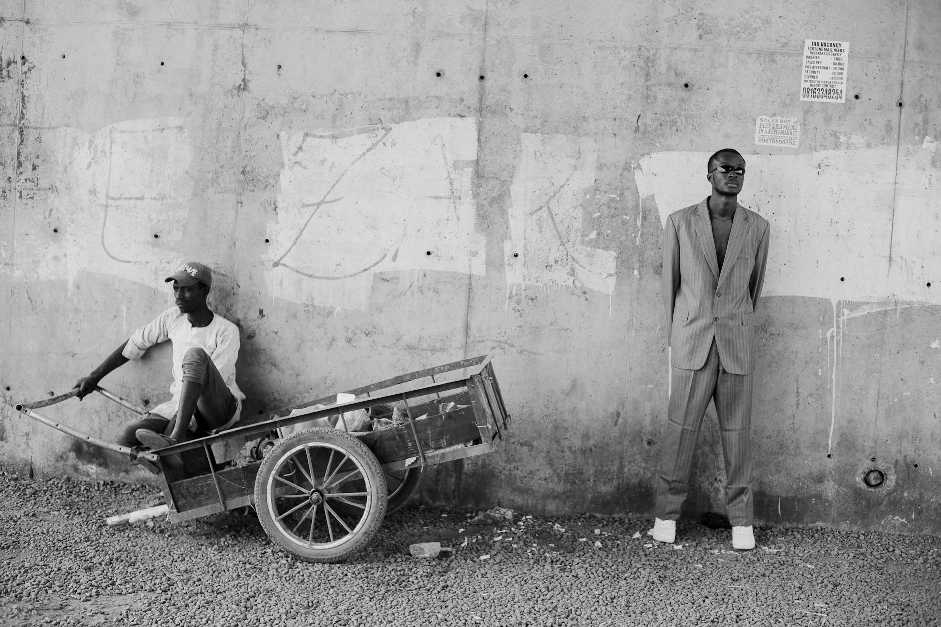 Two men pose in contrasting styles beside a concrete wall in an urban setting.