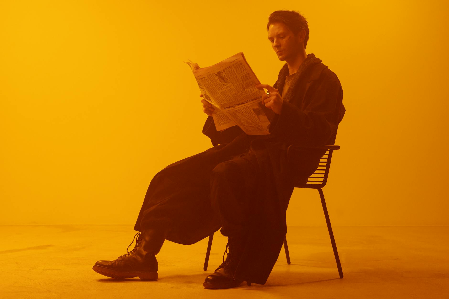 A man sits in a chair, focused on reading a newspaper in a warm, sepia-toned studio setting.