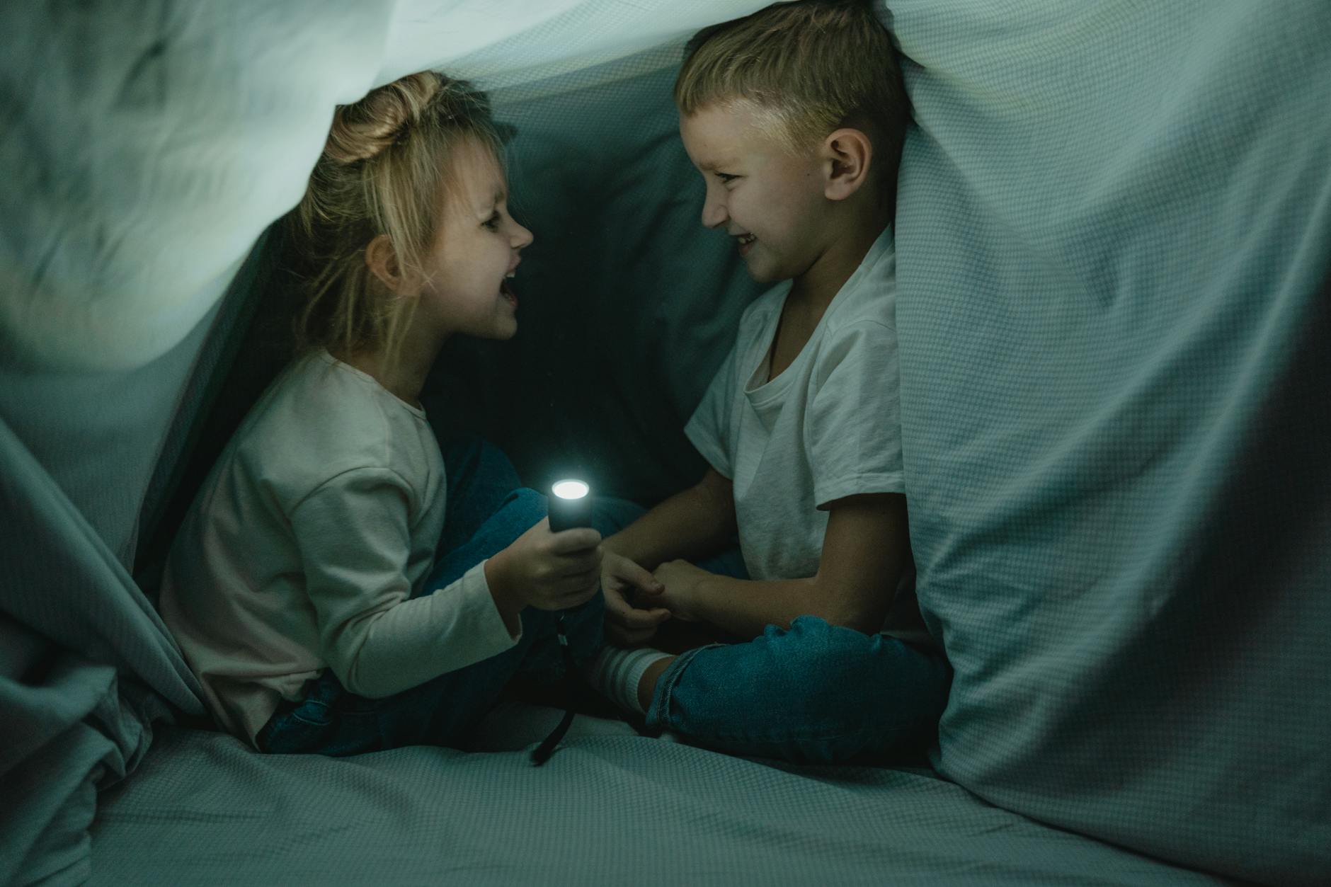 Two children giggle in a cozy blanket fort, sharing stories in flashlight glow.