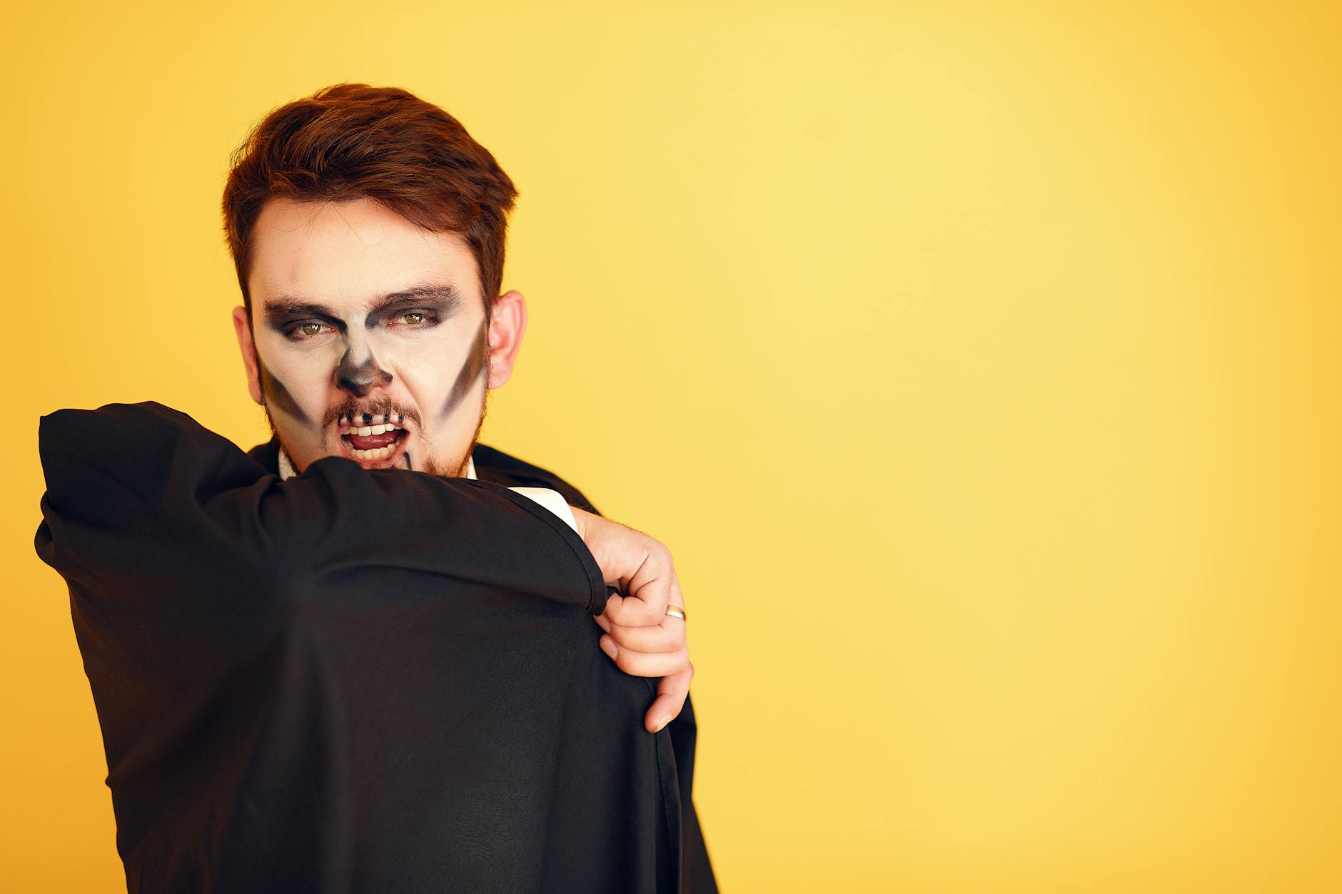 Spooky portrait of a man in skull face paint posing with a dark cloak against a yellow backdrop.