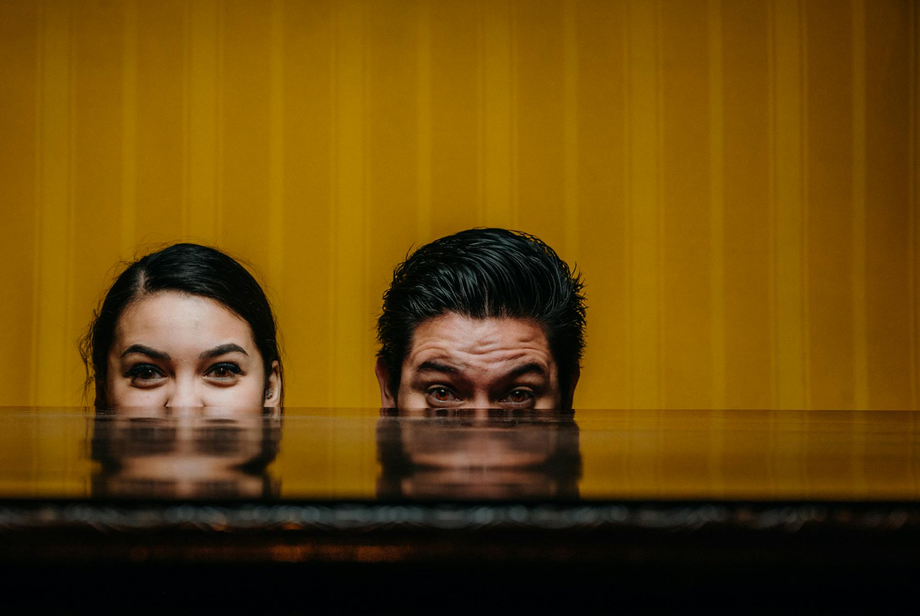 A creative portrait of a man and woman playfully peeking over a reflective surface, set against a vibrant yellow background.