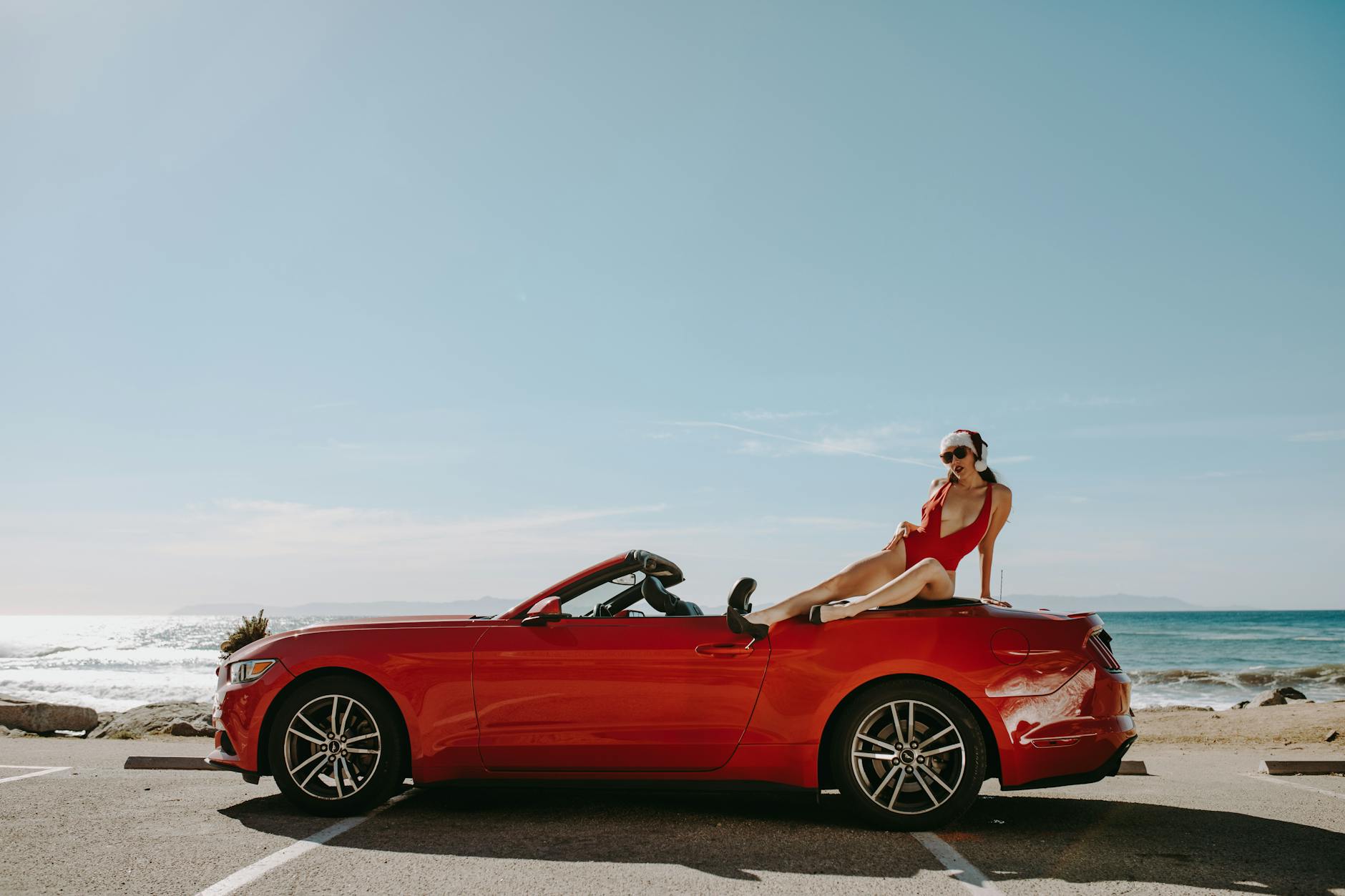A woman in a red swimsuit lounging on a red convertible by the ocean, enjoying a sunny day.