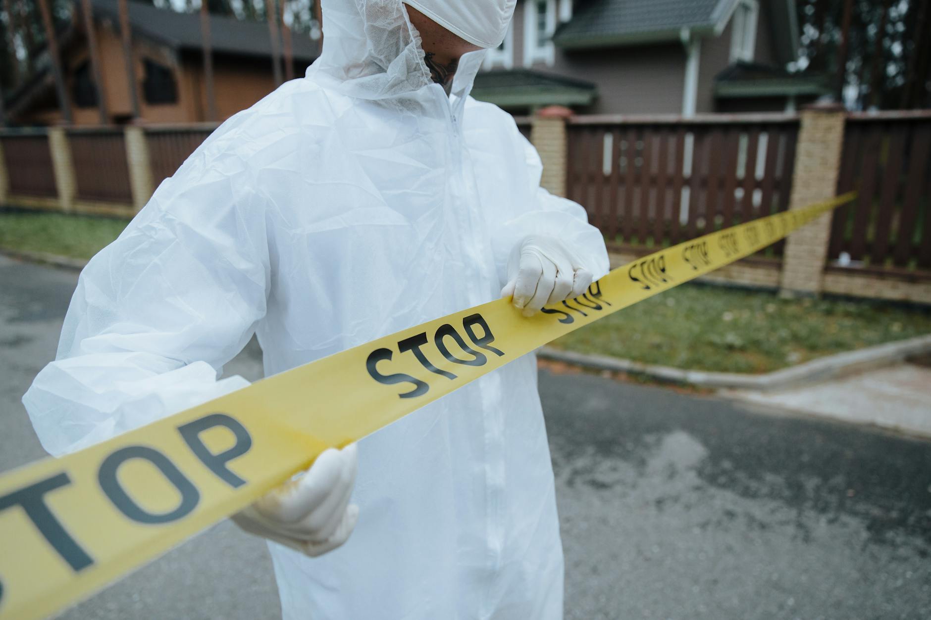 A person wearing a protective suit, handling yellow tape with 'STOP' in an outdoor urban setting.