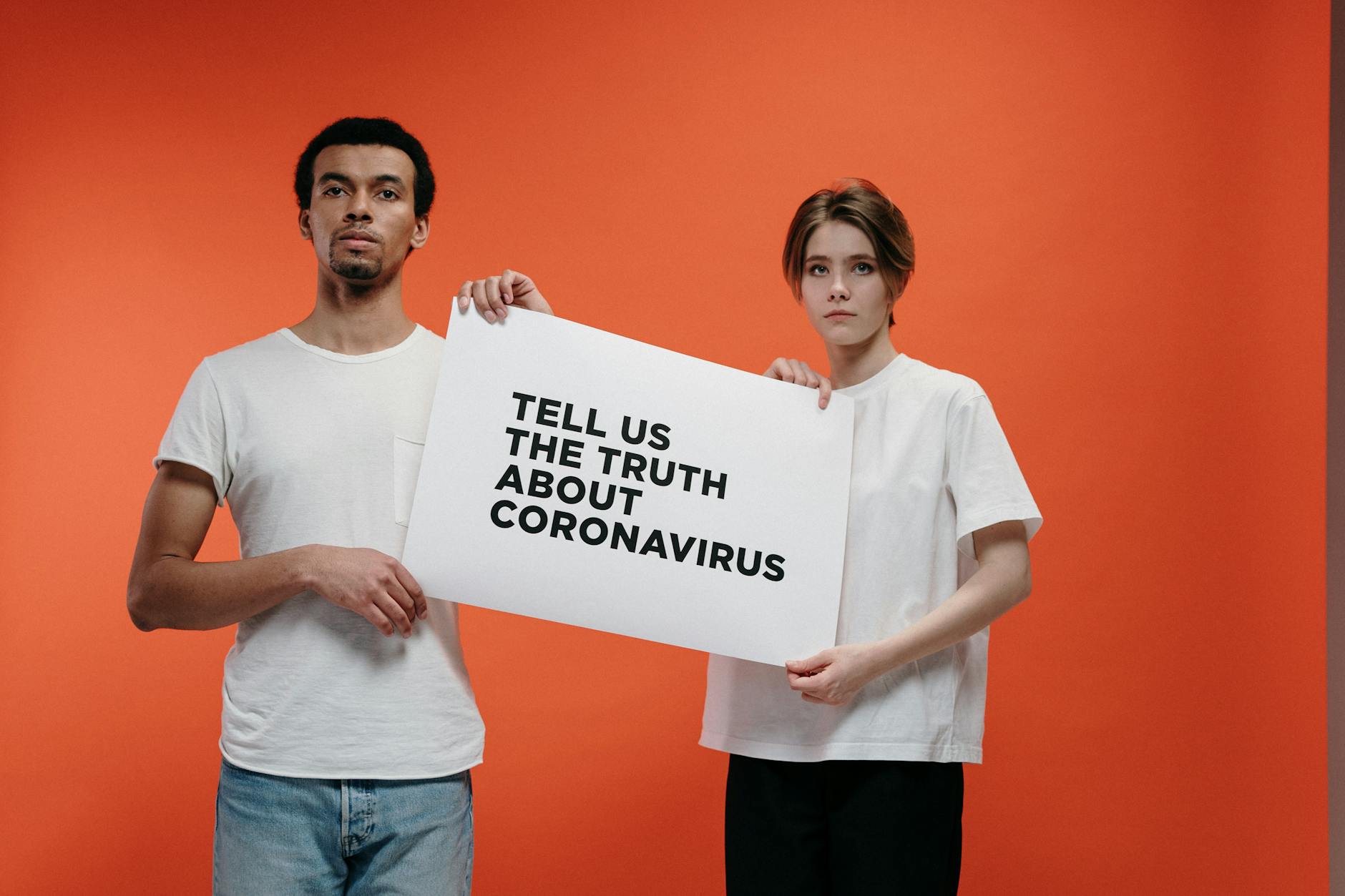 Two people holding a 'Tell Us The Truth About Coronavirus' sign against a vibrant orange background.