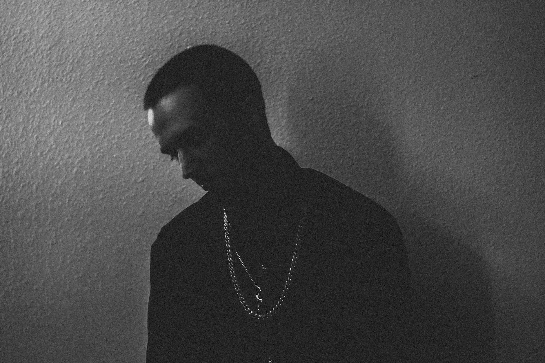 A moody black and white portrait of a man wearing chains against a textured wall.