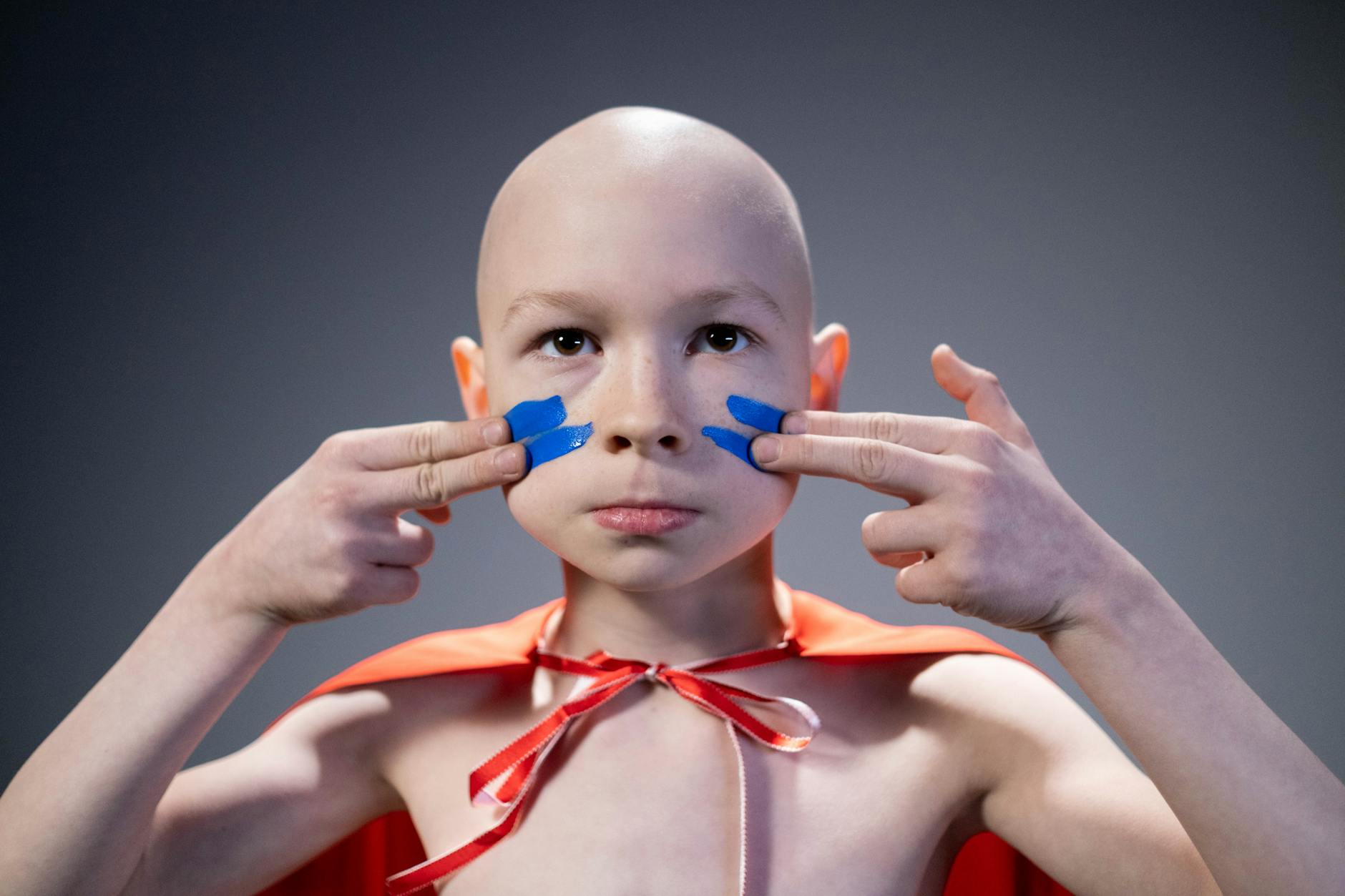Portrait of a bald child wearing a red cape and blue face paint, embodying strength and courage.