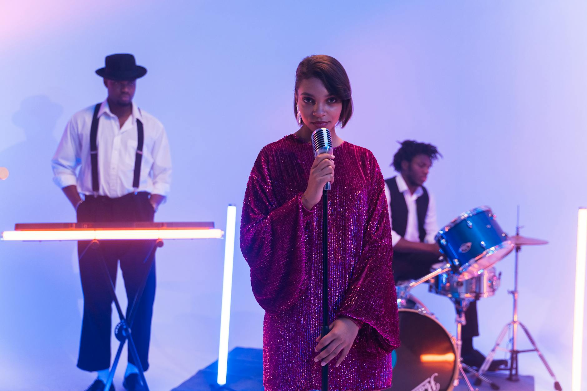 A vibrant band performing indoors with a singer, keyboardist, and drummer under colorful lights.