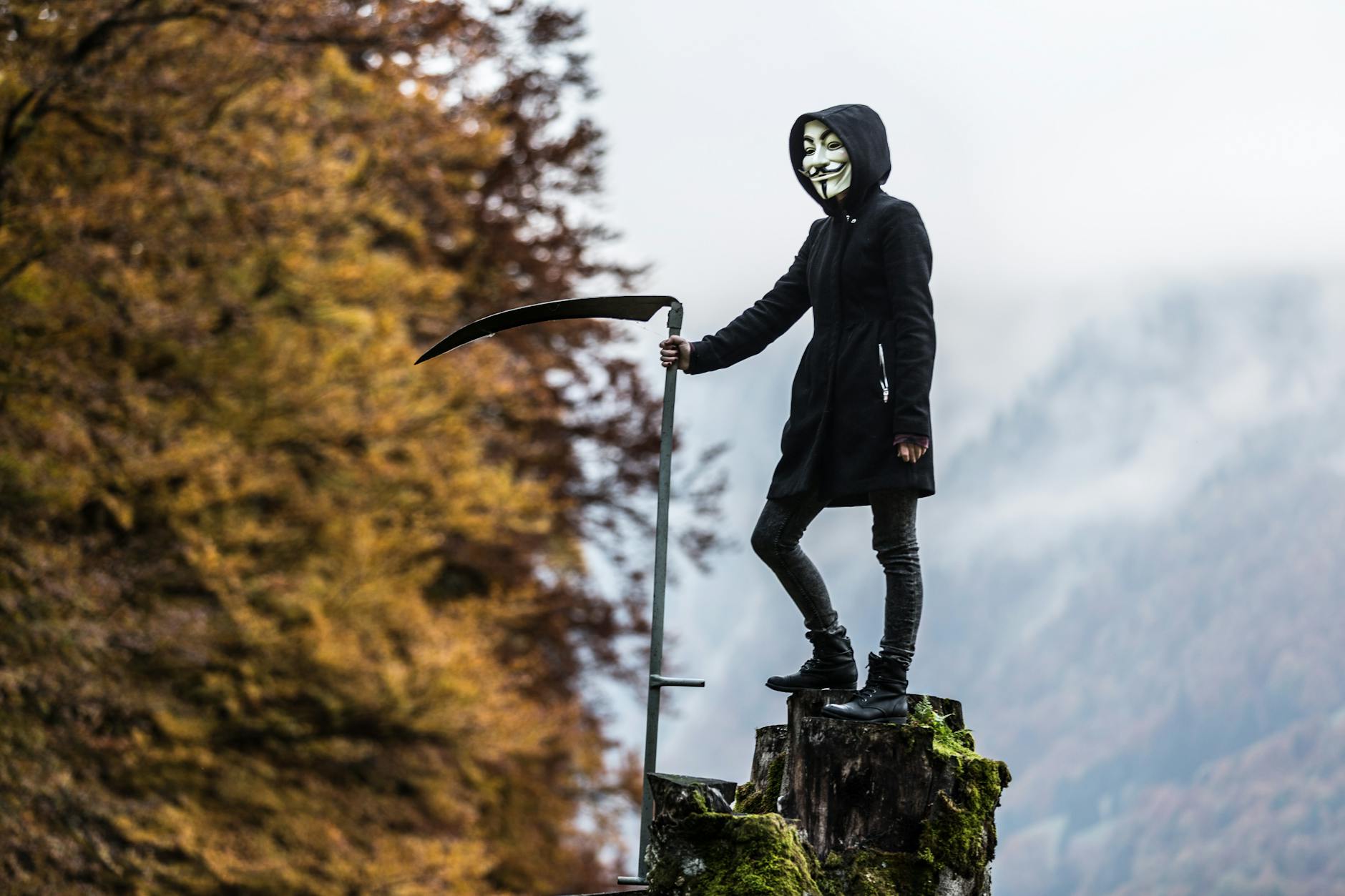 A masked figure holding a scythe stands on a tree stump amidst autumn foliage in a misty setting.